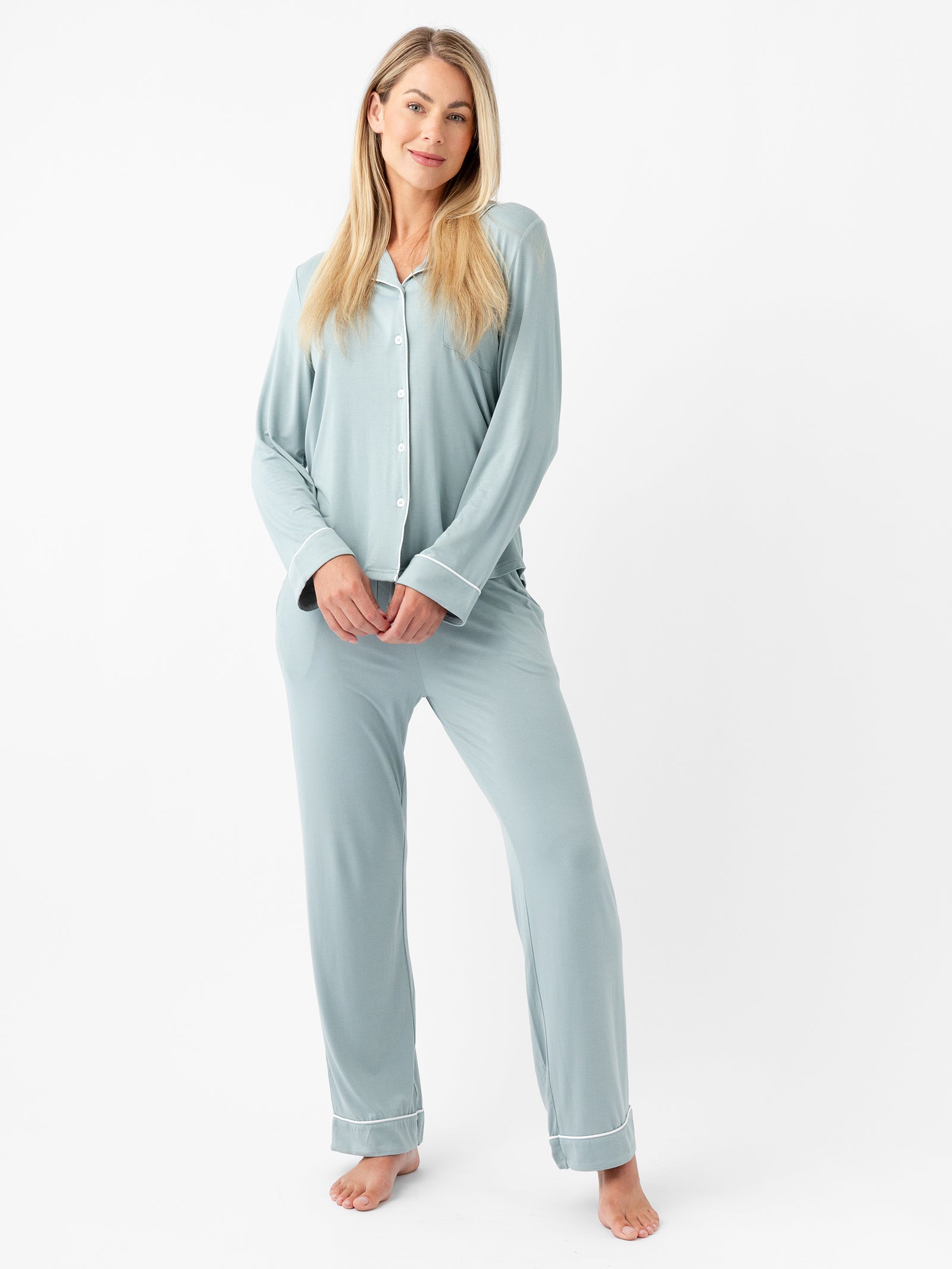 A woman stands on a white background wearing the Women's Long Sleeve Bamboo Pajama Top in Stretch-Knit from HIDE. The light blue pajama set features buttons down the front and white trim on the cuffs and pockets. She appears relaxed with her hands resting at her sides, and she is barefoot. 