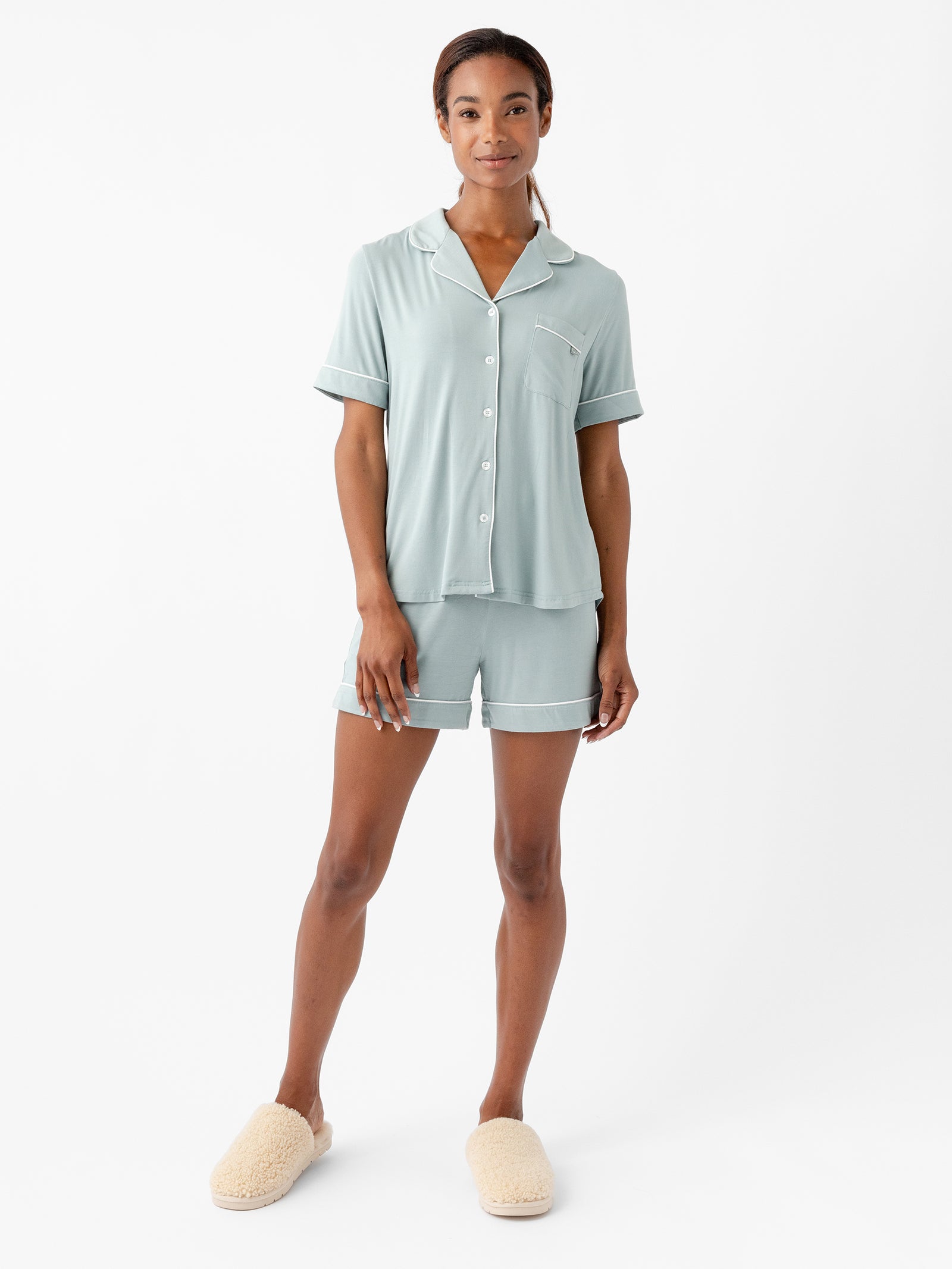 A person stands against a plain white background wearing a light green Women's Short Sleeve Bamboo Pajama Top in Stretch-Knit by HIDE, featuring white trim. The pajama set includes a short-sleeve button-up top and matching shorts. They complete the look with beige fluffy slippers, maintaining a relaxed posture and slight smile. 