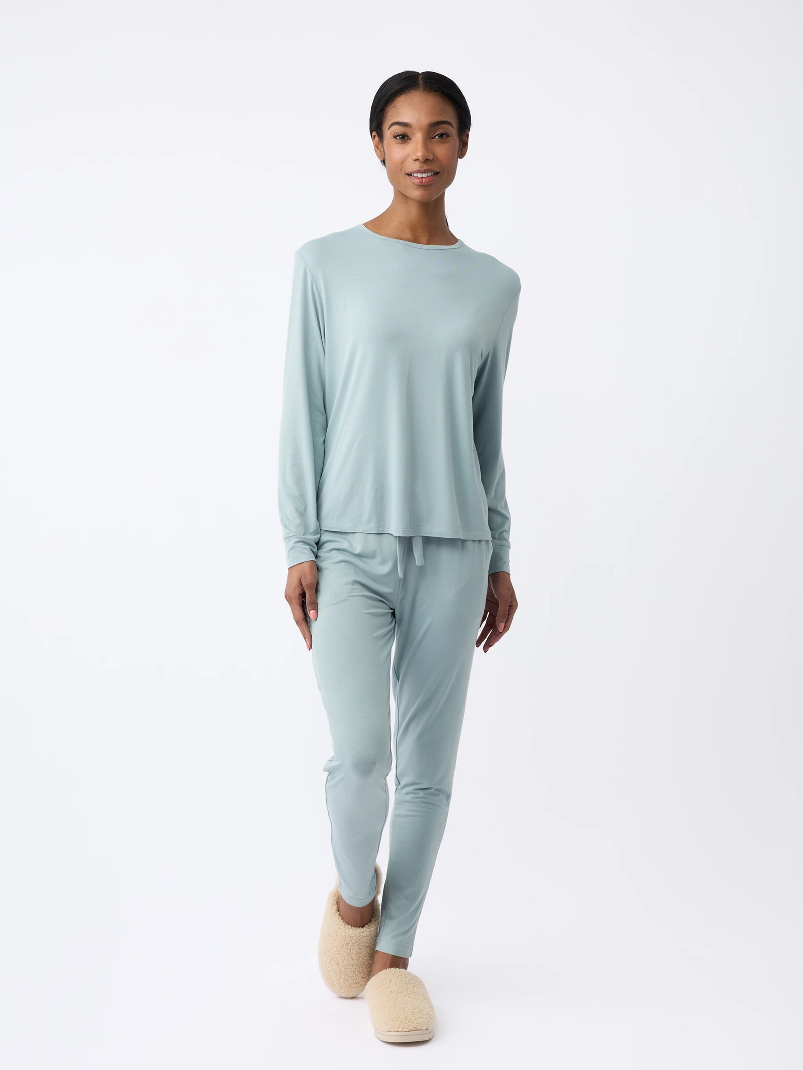 A woman wearing Cozy Earth's Women's Stretch Knit Bamboo Long Sleeve Lounge Tee in light blue with matching pants stands against a plain white background, sporting plush beige slippers and gazing forward with a relaxed expression. 