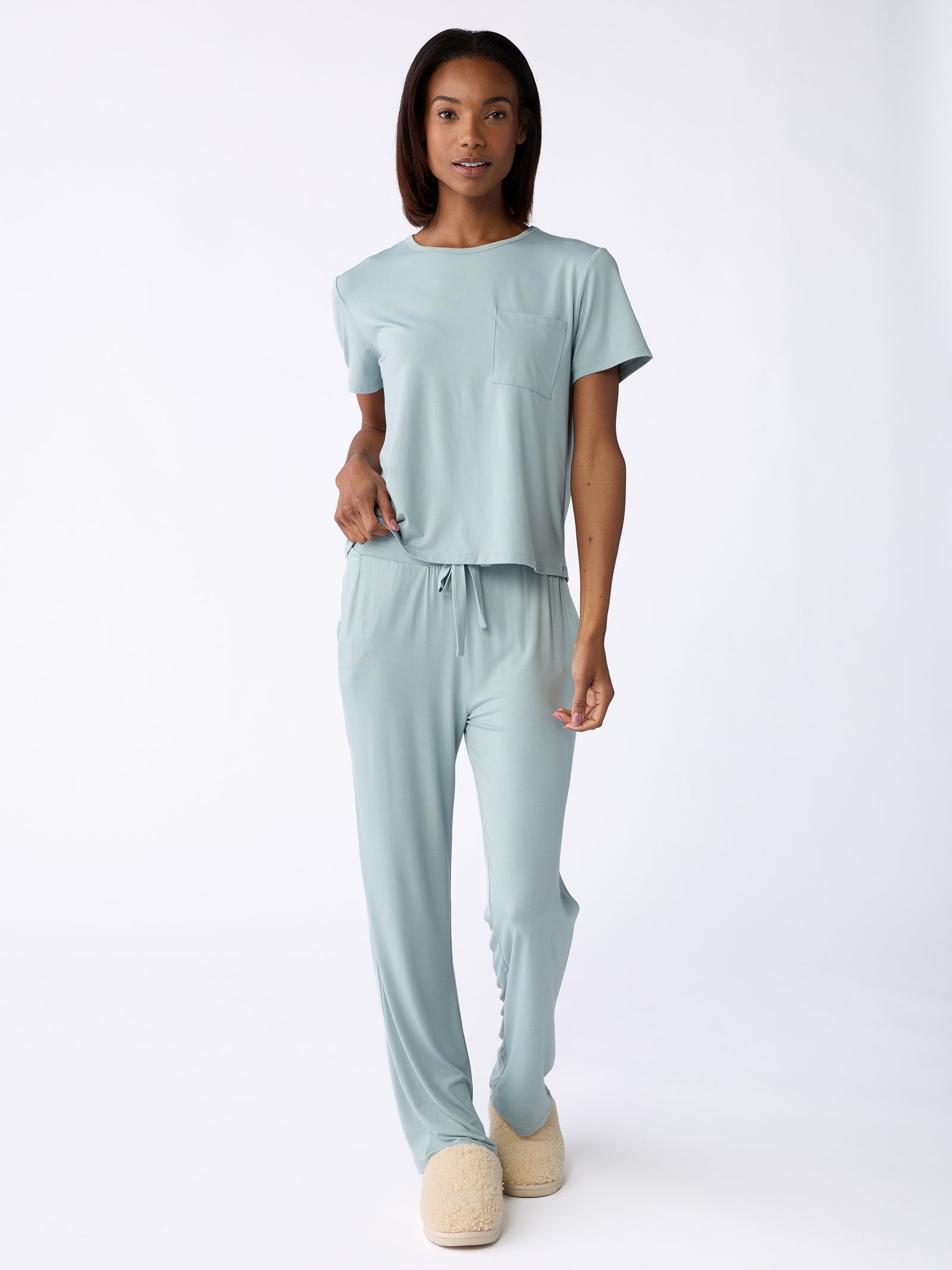 A person wearing the Stretch Knit Bamboo Pant by Cozy Earth, featuring a light blue pajama set with a short-sleeve top and drawstring pants, stands against a white background. They are also wearing light tan slippers. 