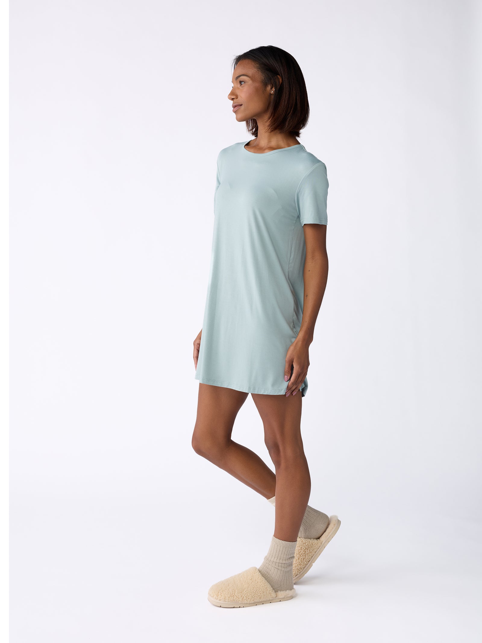 A woman stands in a relaxed pose with a subtle smile, wearing the Women's Bamboo Stretch Knit Sleep Dress by Cozy Earth. She pairs the light blue dress with beige socks and tan slippers, set against a plain white background. 