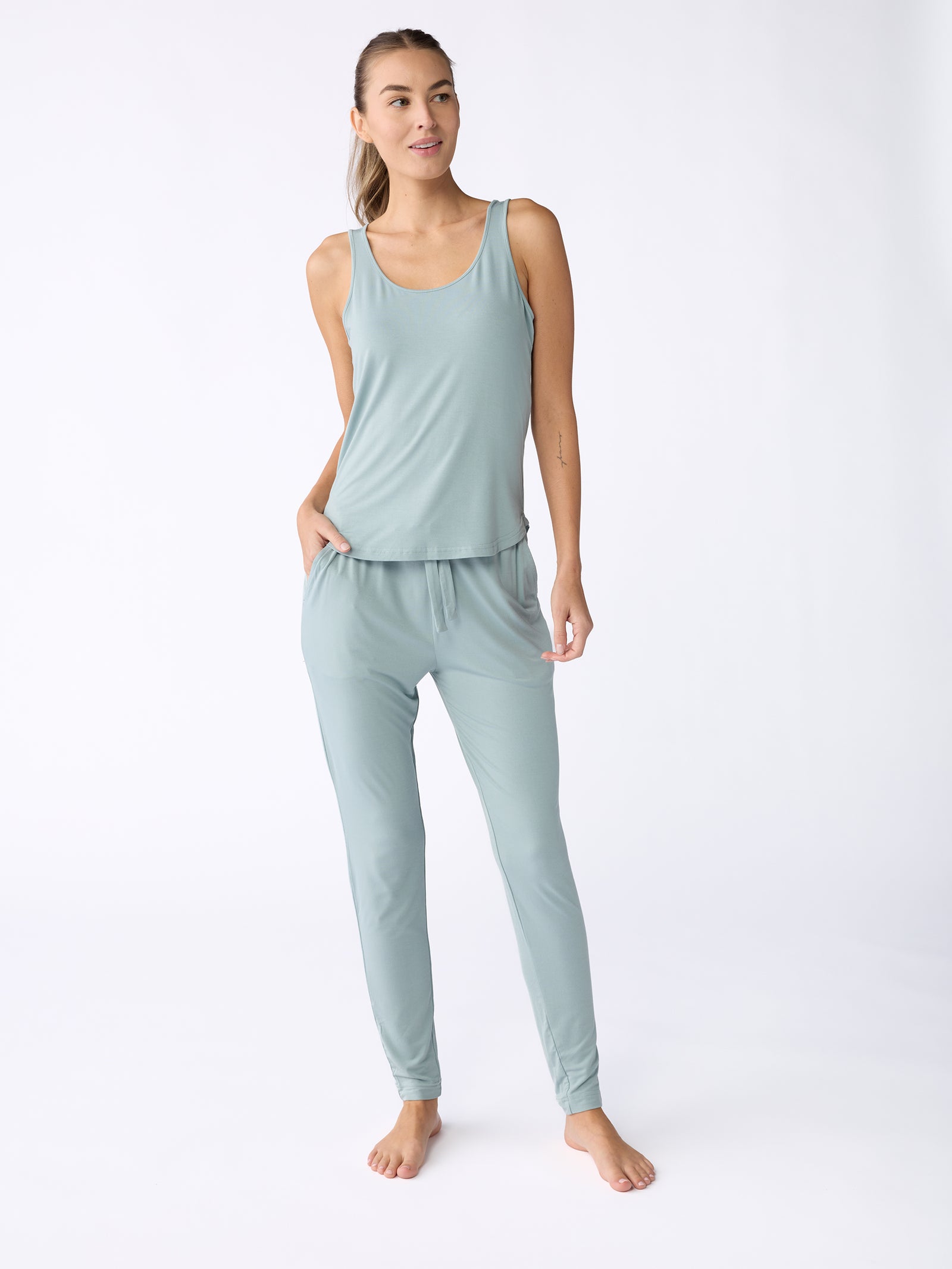 A woman is posed casually, donning a light teal sleeveless top paired with Cozy Earth's Stretch Knit Bamboo Tapered Pant. Barefoot against a white backdrop, she wears a subtle smile while gazing to the side. 