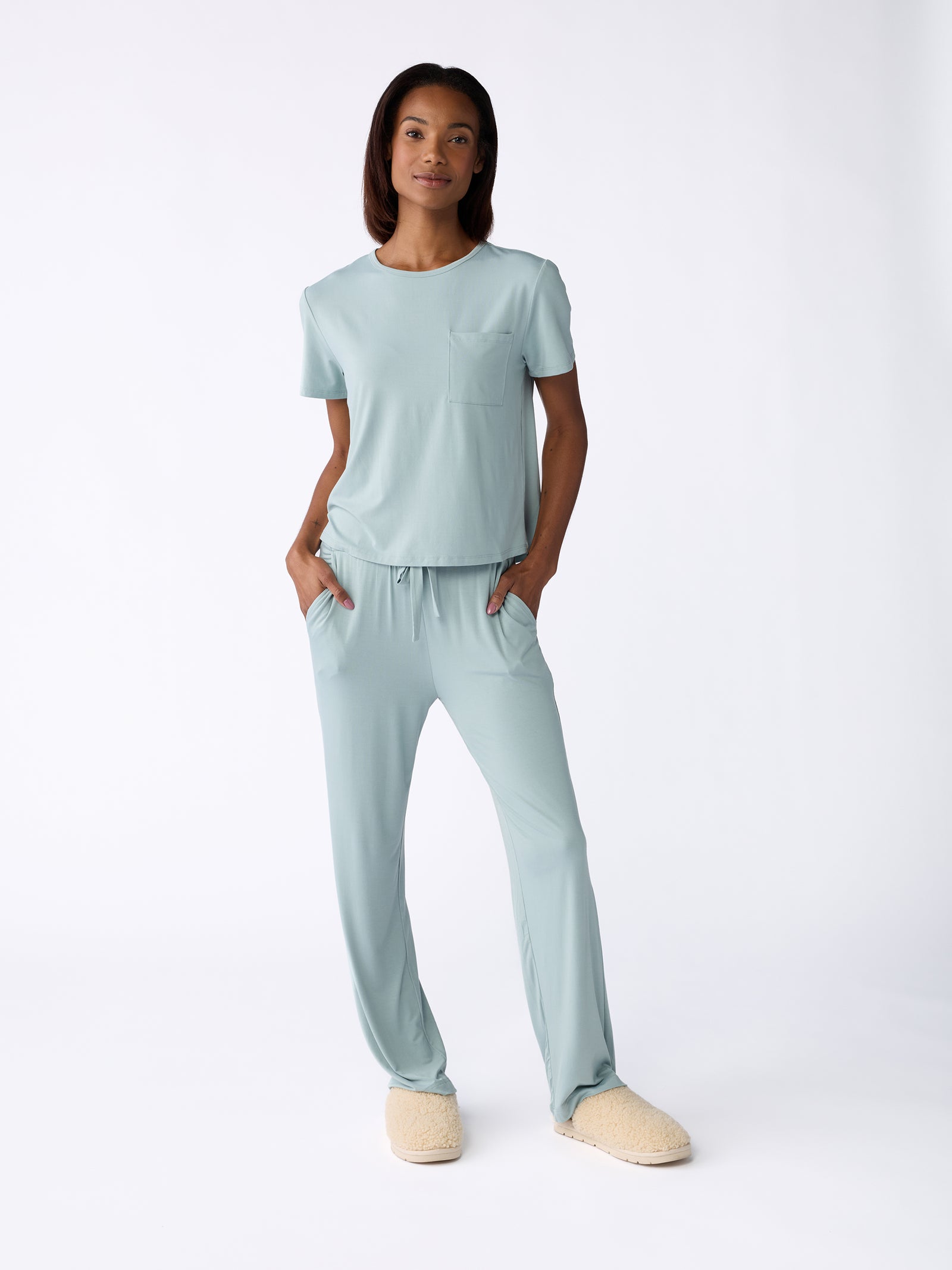 Dressed in a relaxed, light blue ensemble from Cozy Earth's Women's Stretch-Knit Bamboo Lounge Tee collection, the person exudes confidence. They have their hands casually tucked into pockets and complement their outfit with cozy, cream-colored slippers, all against a plain white background. 