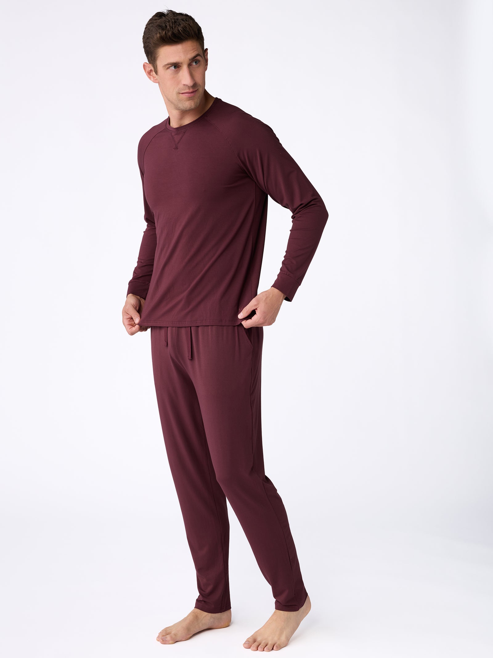 A person wearing the Men's Stretch-Knit Bamboo Long Sleeve by Cozy Earth in maroon, along with matching pants, stands barefoot on a white background, looking to the side. 