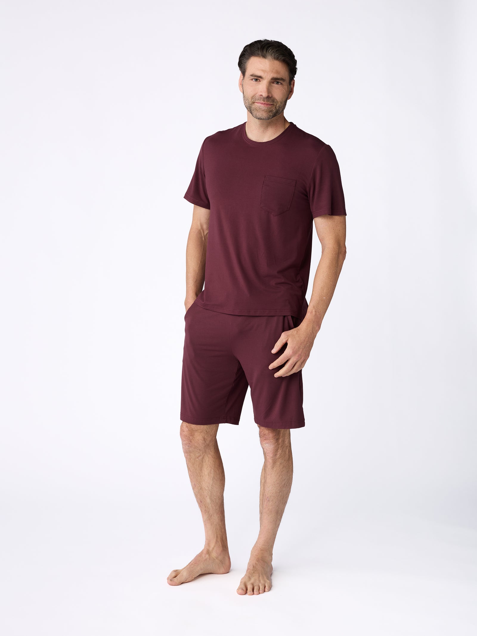 A man stands barefoot against a plain white background, dressed in a Cozy Earth burgundy men's stretch-knit bamboo pajama short set. He has short dark hair and a light beard, smiling with one hand in his pocket. 