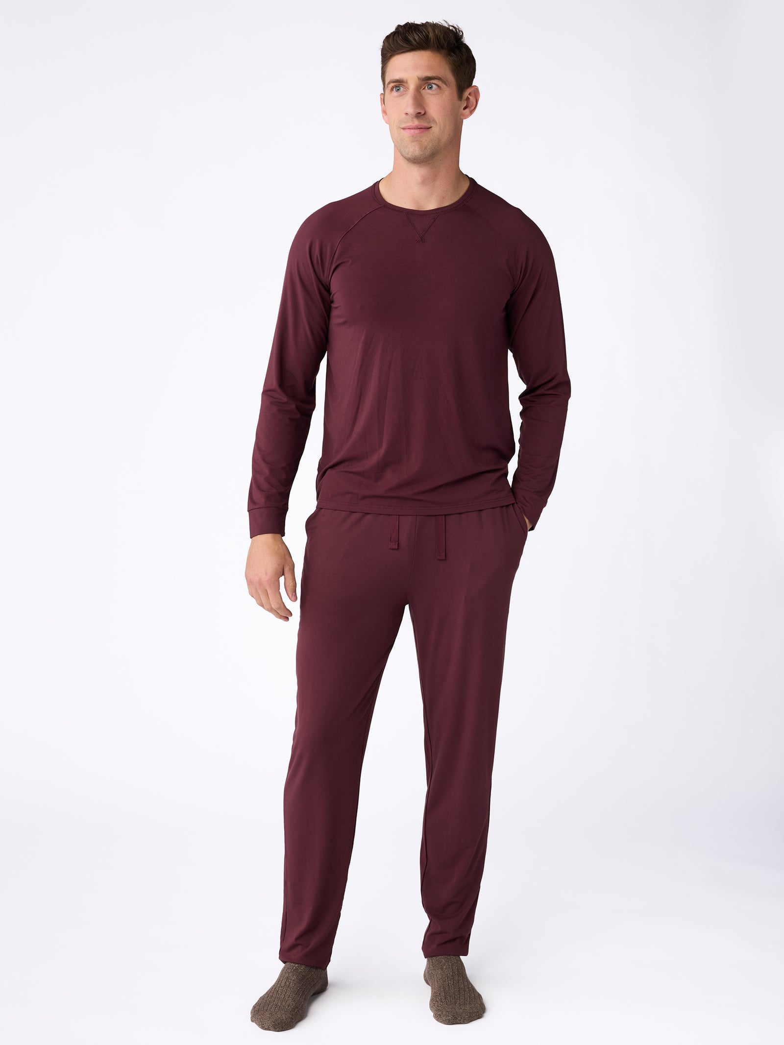 A man is standing on a white background wearing a long-sleeved maroon pajama set, including the Men's Stretch-Knit Bamboo Pajama Pant from Cozy Earth, and gray socks. 