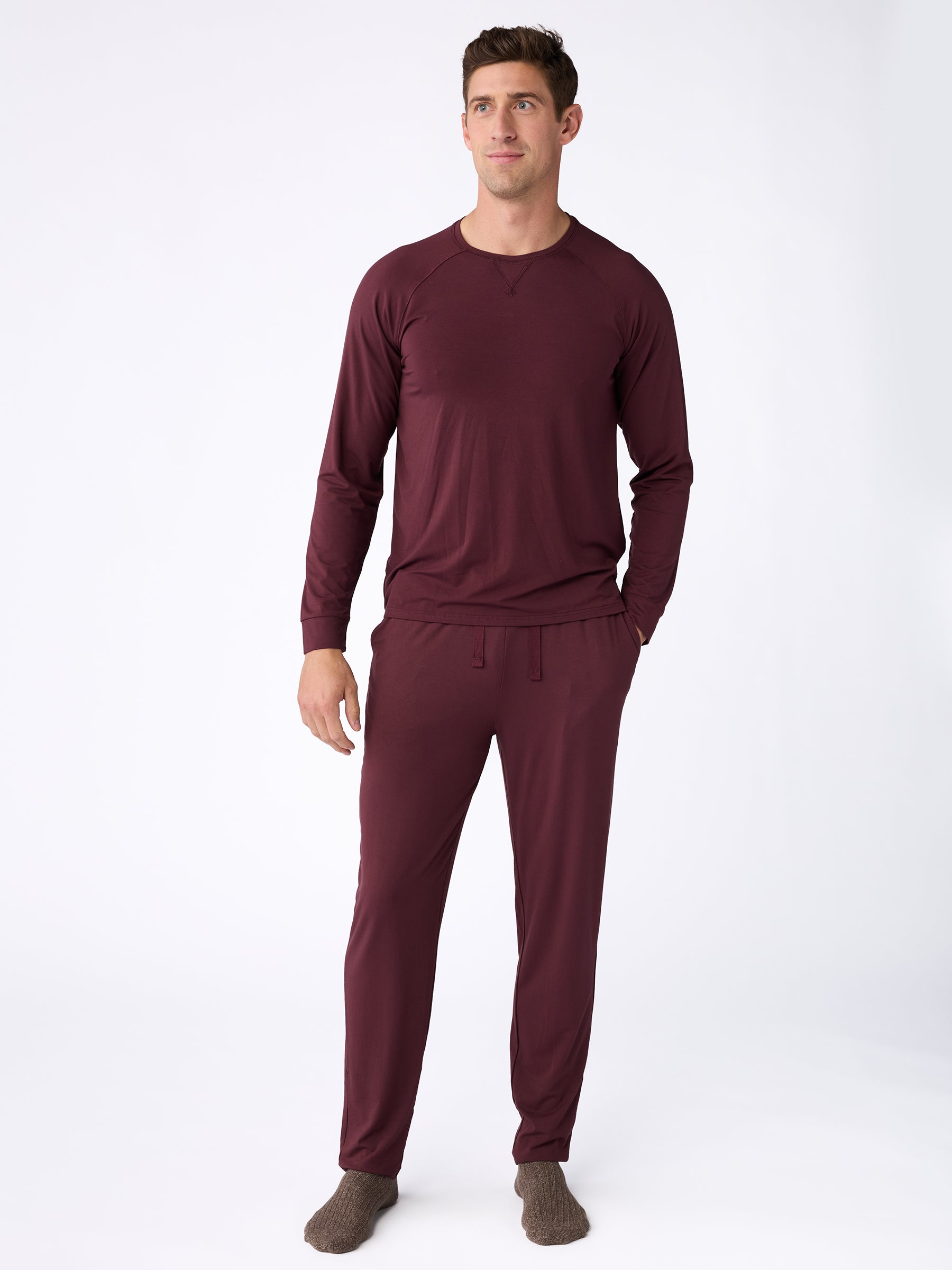 A man is standing on a white background wearing a long-sleeved maroon pajama set, including the Men's Stretch-Knit Bamboo Pajama Pant from Cozy Earth, and gray socks. |Color:Burgundy