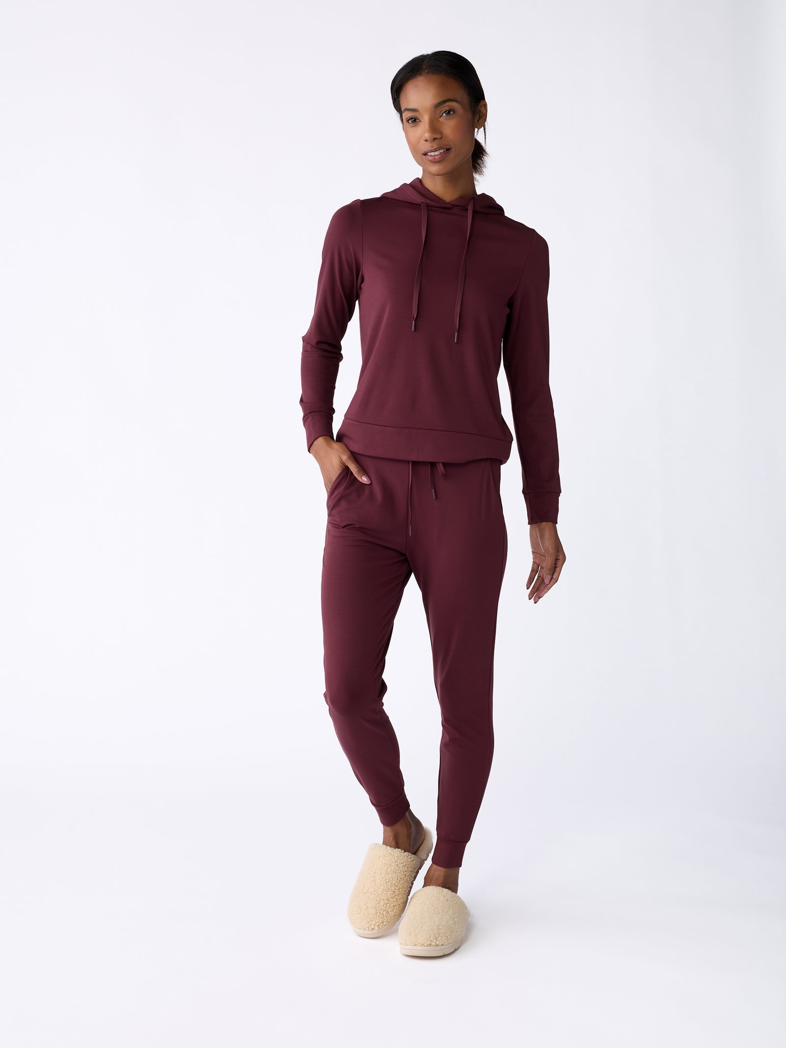 A person wearing the Women's Ultra-Soft Bamboo Hoodie from Cozy Earth, in burgundy, is casually standing against a plain white background. They're also styled with matching joggers and beige fluffy slippers, with one hand in their pocket and a relaxed expression. 