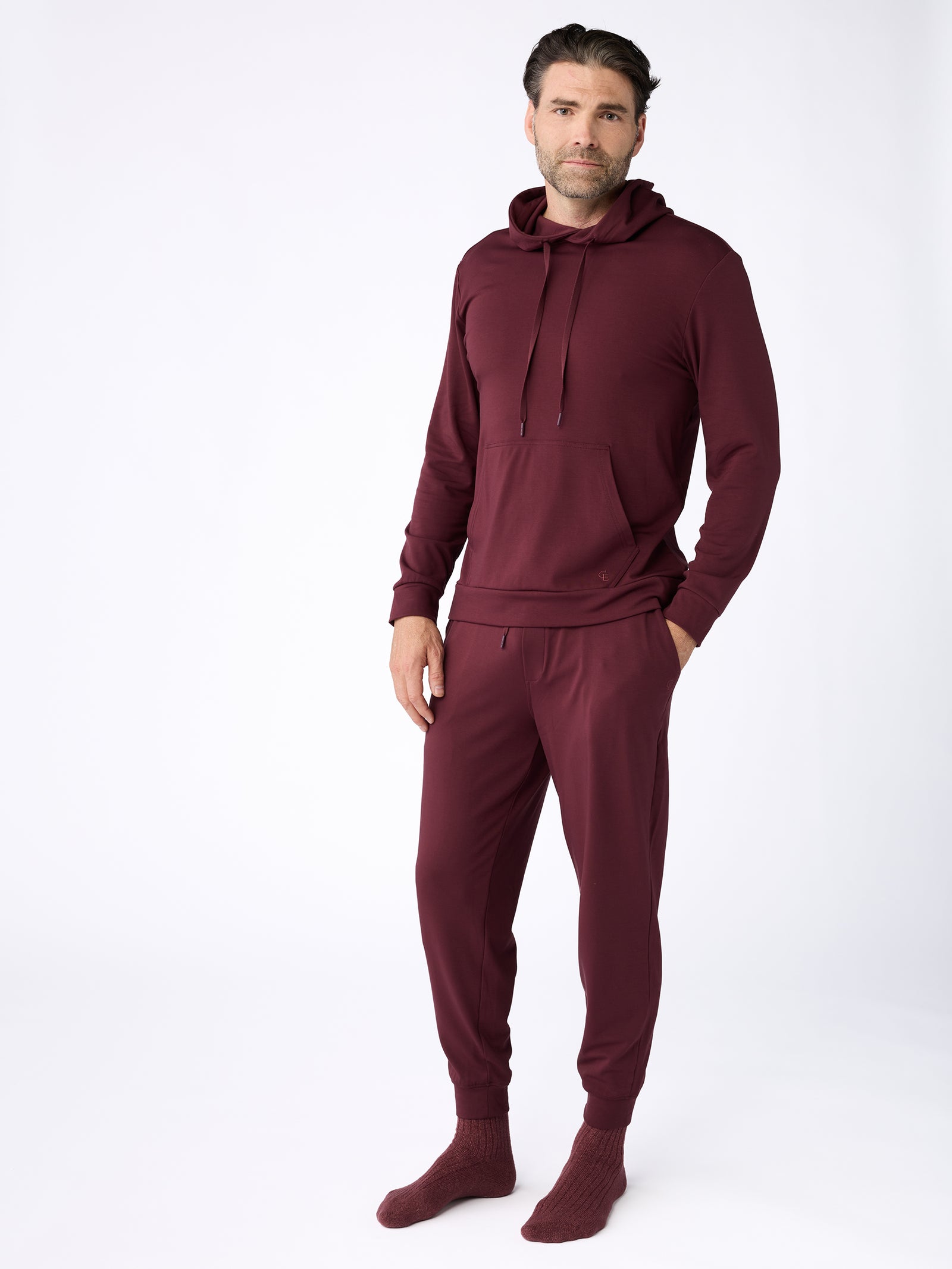 A man stands against a plain white background wearing the Men's Ultra-Soft Bamboo Hoodie by Cozy Earth in maroon, paired with matching joggers. He has a relaxed posture, with one hand in his pocket. 