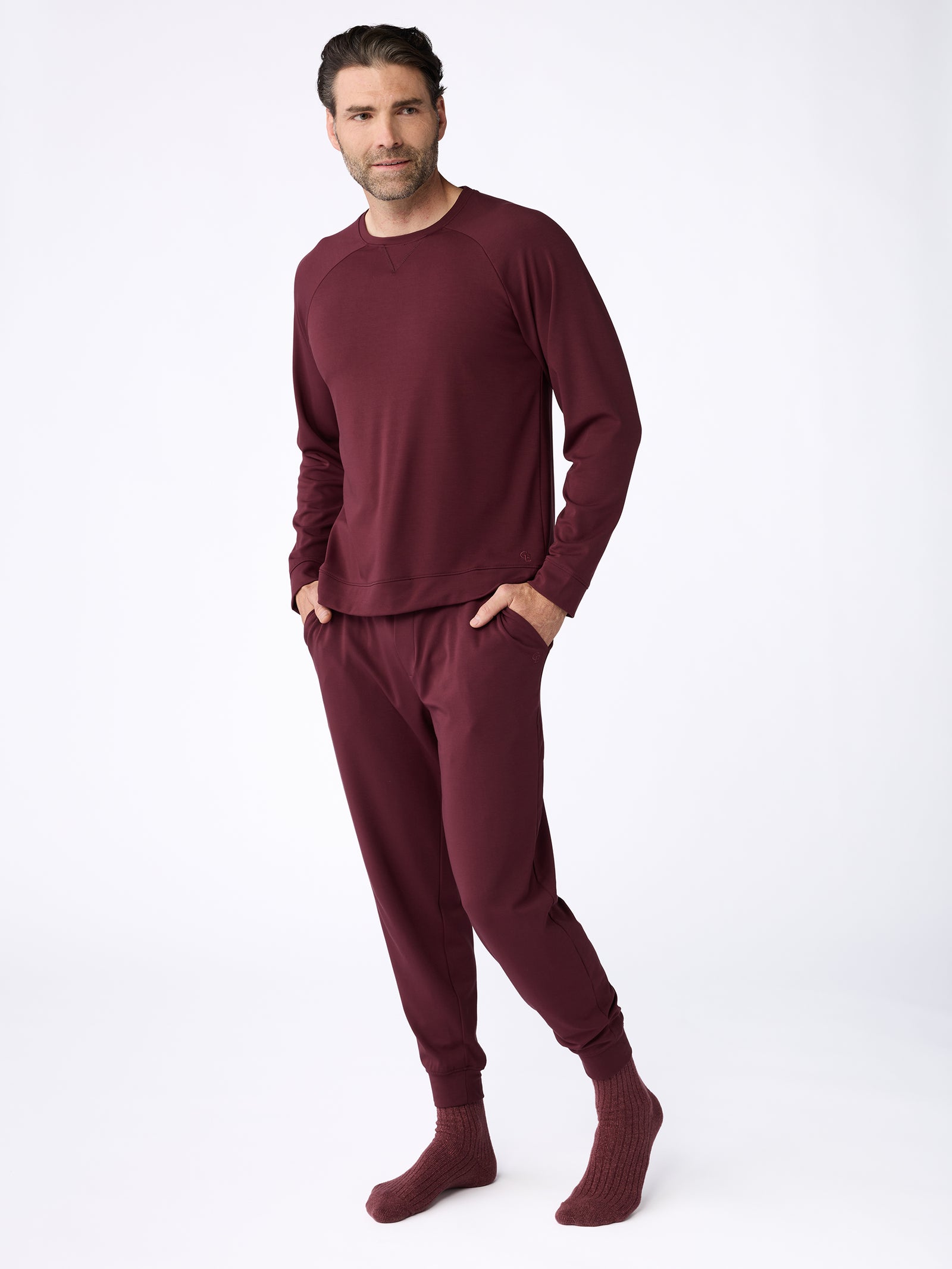 A person is seen standing with their hands in the pockets of a matching maroon loungewear set, featuring Cozy Earth's Men's Ultra-Soft Bamboo Pullover Crew and coordinating pants. They are also wearing maroon socks. The backdrop is simple and light-colored. 