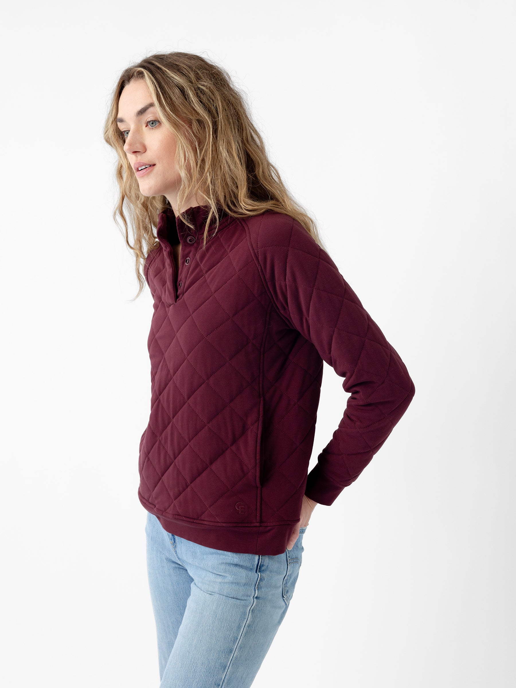 A woman with long wavy hair wearing the maroon Women's Ultra-Soft Bamboo Quilted Snap Pullover from Cozy Earth and light blue jeans stands against a plain white background. She looks to her right with a slight smile on her face, with her hands in her pockets. |Color:Burgundy