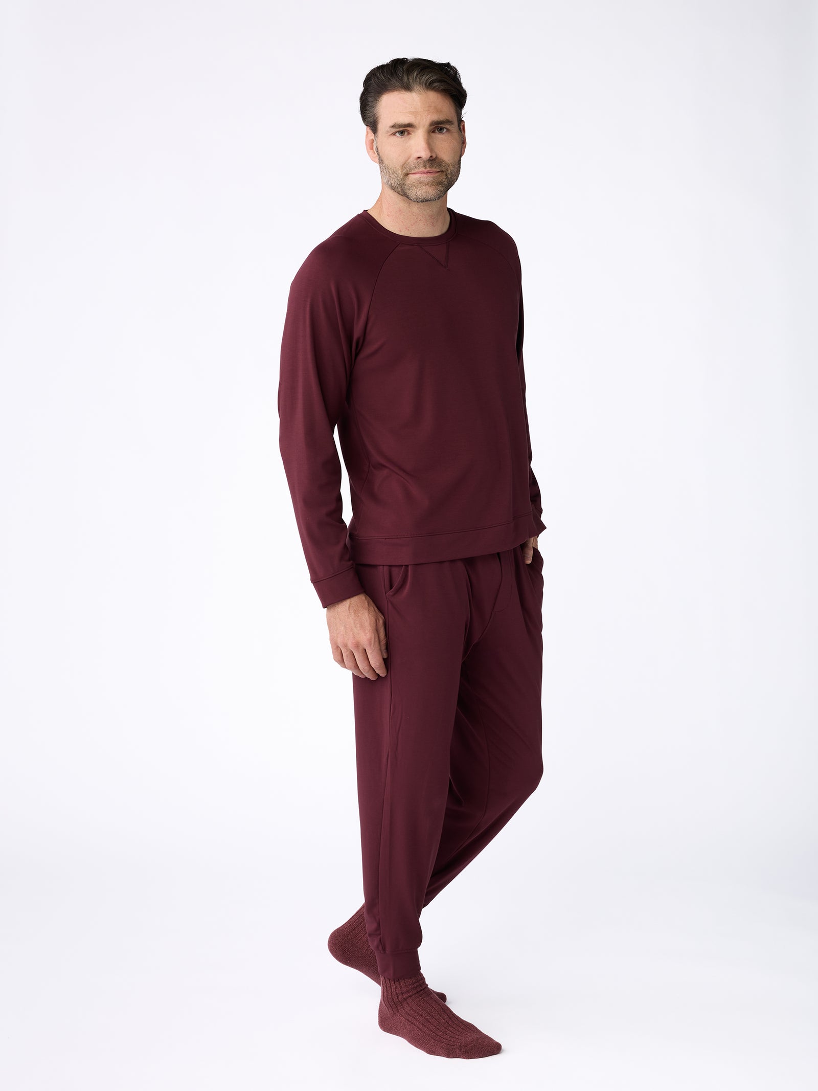 A man dressed in the Cozy Earth Men's Ultra-Soft Bamboo Pullover Crew and matching pants poses against a plain white background. With dark hair and a relaxed expression, he gazes at the camera while standing barefoot. 