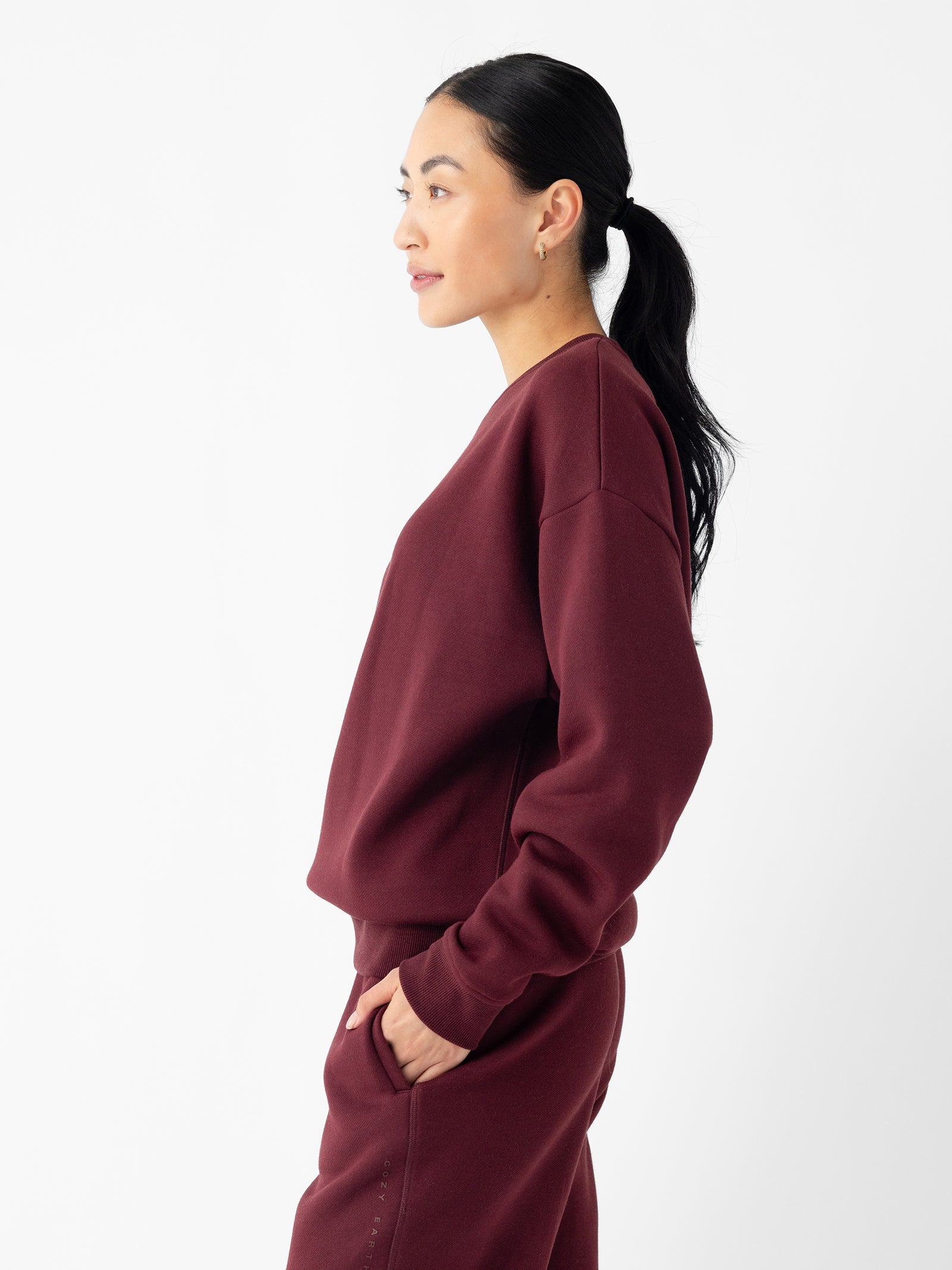 A person with dark hair tied back in a ponytail is standing in profile against a white background. They are wearing a matching maroon Women's CityScape Crewneck top and pants from Cozy Earth, and have their right hand in the pant pocket while looking slightly ahead. 