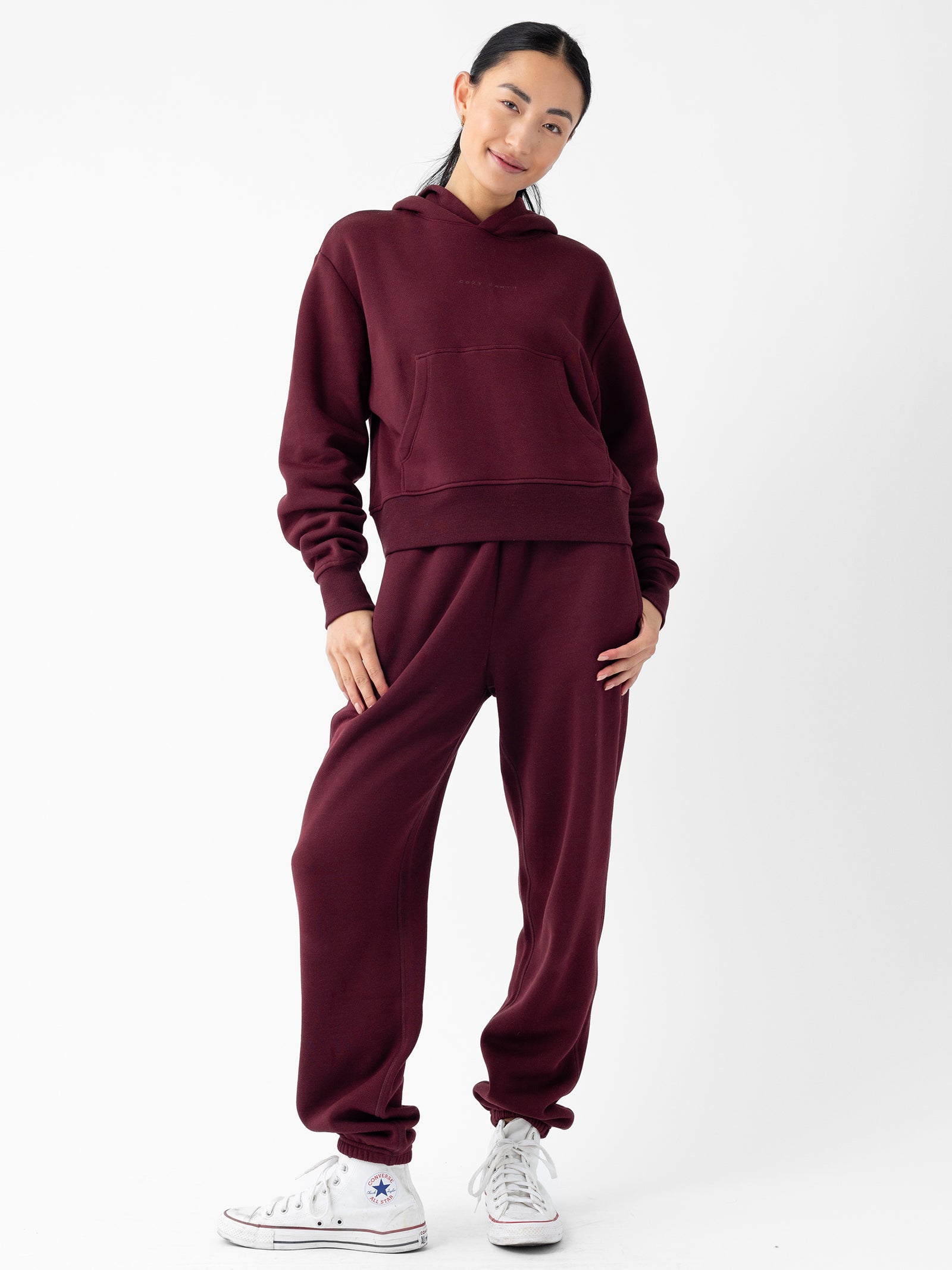 A young woman stands against a white background, smiling with her hands in her pockets. She is wearing a Cozy Earth Women's CityScape Cropped Hoodie in burgundy, matching sweatpants, and white sneakers. Her hair is tied back in a ponytail. 