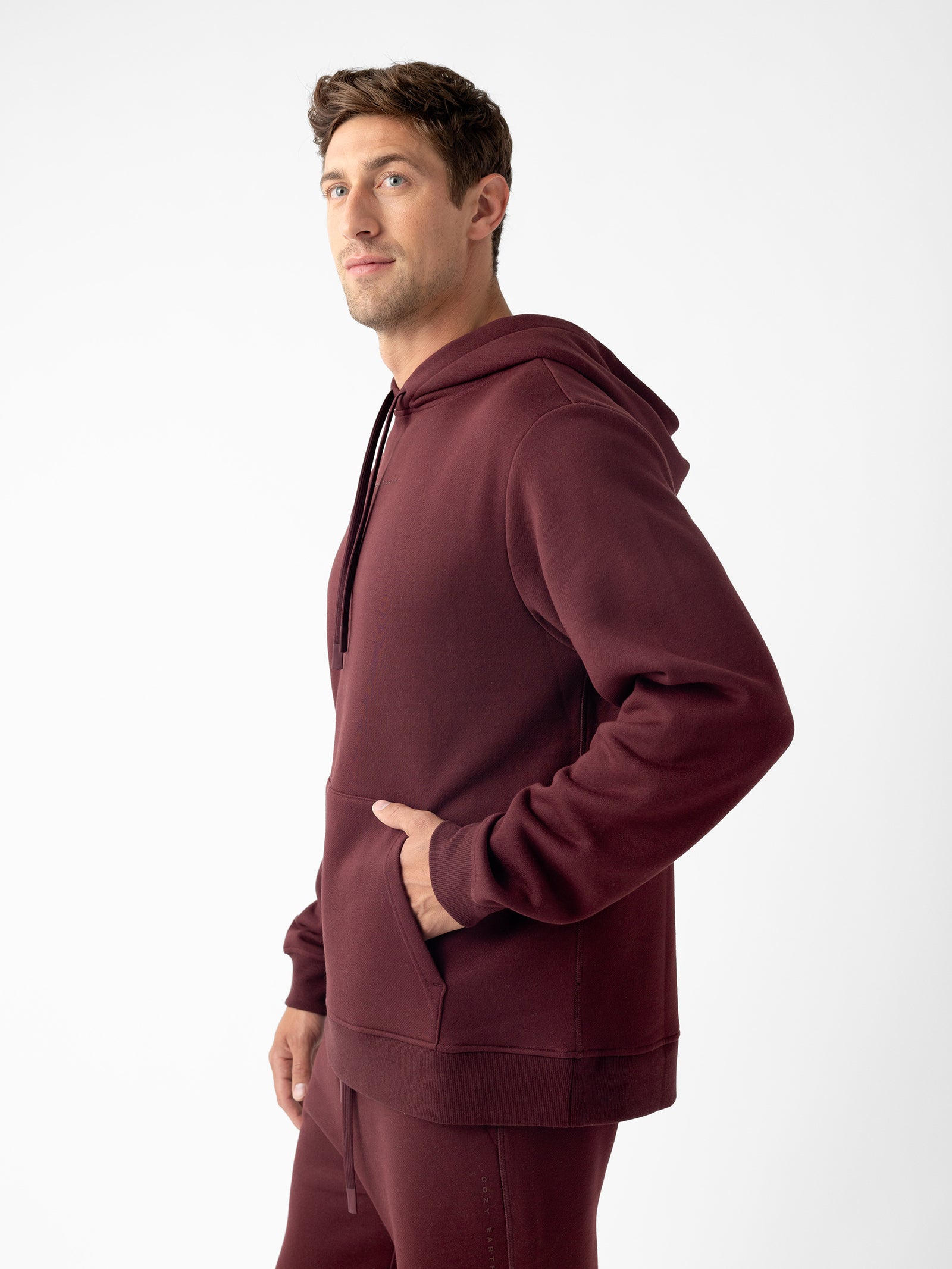 A man wearing a Cozy Earth Men's CityScape Hoodie in maroon, along with matching pants, stands against a plain white background. He has his right hand in the front pocket of the hoodie and is looking slightly to the left. 