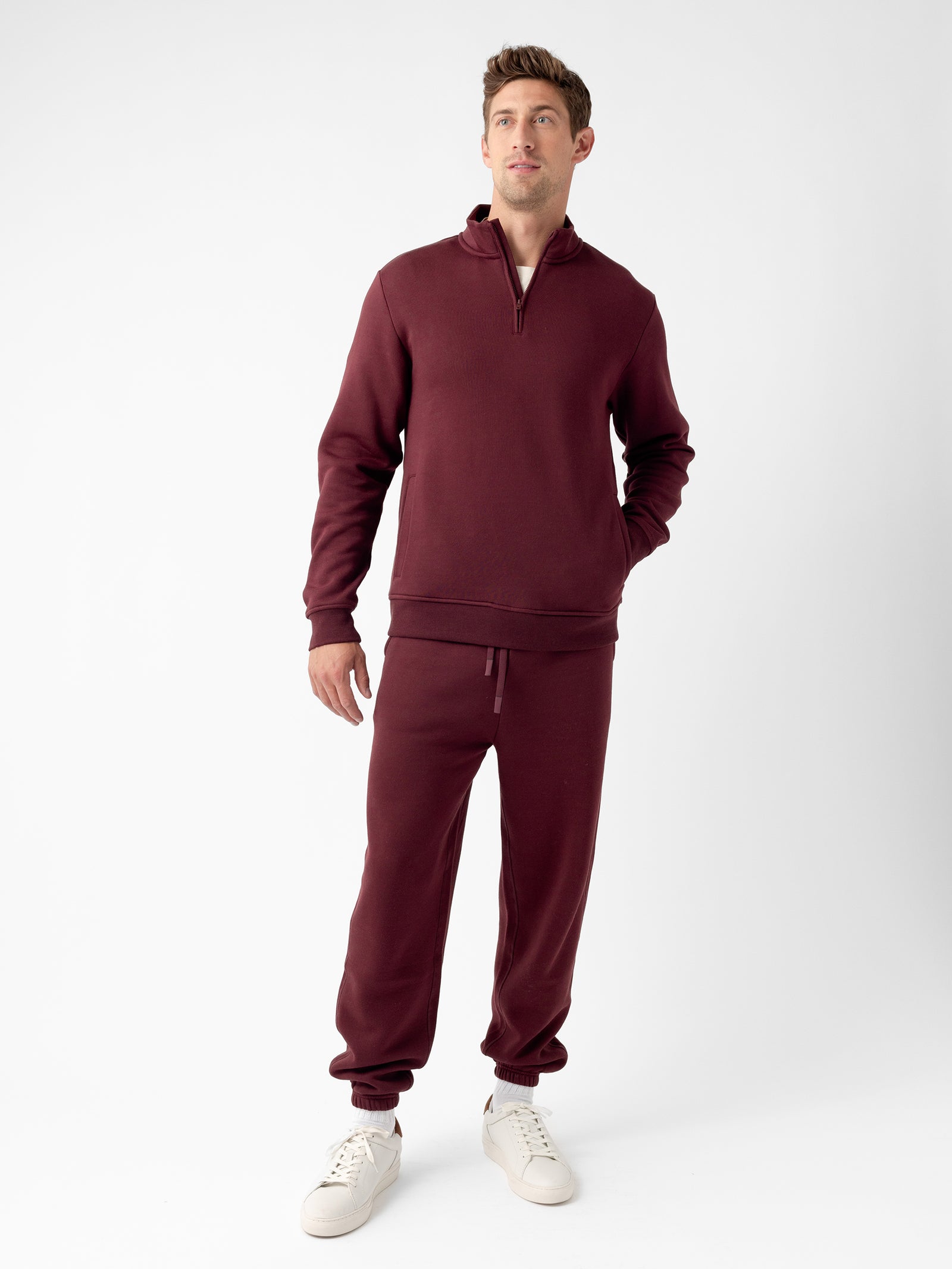 A man stands against a plain white background, wearing a matching maroon Men's CityScape Quarter Zip sweatshirt and jogger pants set by Cozy Earth. He has one hand in his pocket and is looking slightly upwards. He is also wearing white sneakers. 