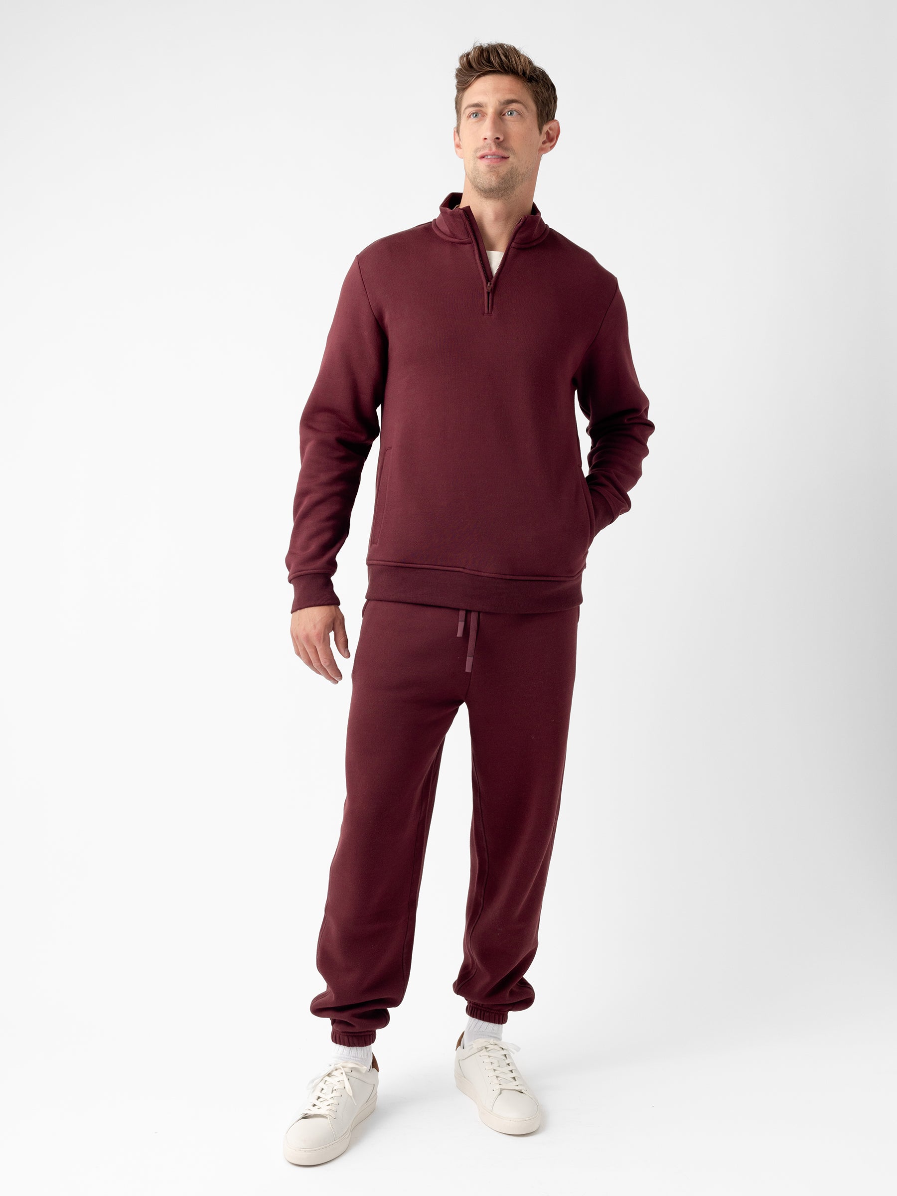 A man stands against a plain white background, wearing a matching maroon Men's CityScape Quarter Zip sweatshirt and jogger pants set by Cozy Earth. He has one hand in his pocket and is looking slightly upwards. He is also wearing white sneakers. |Color:Burgundy