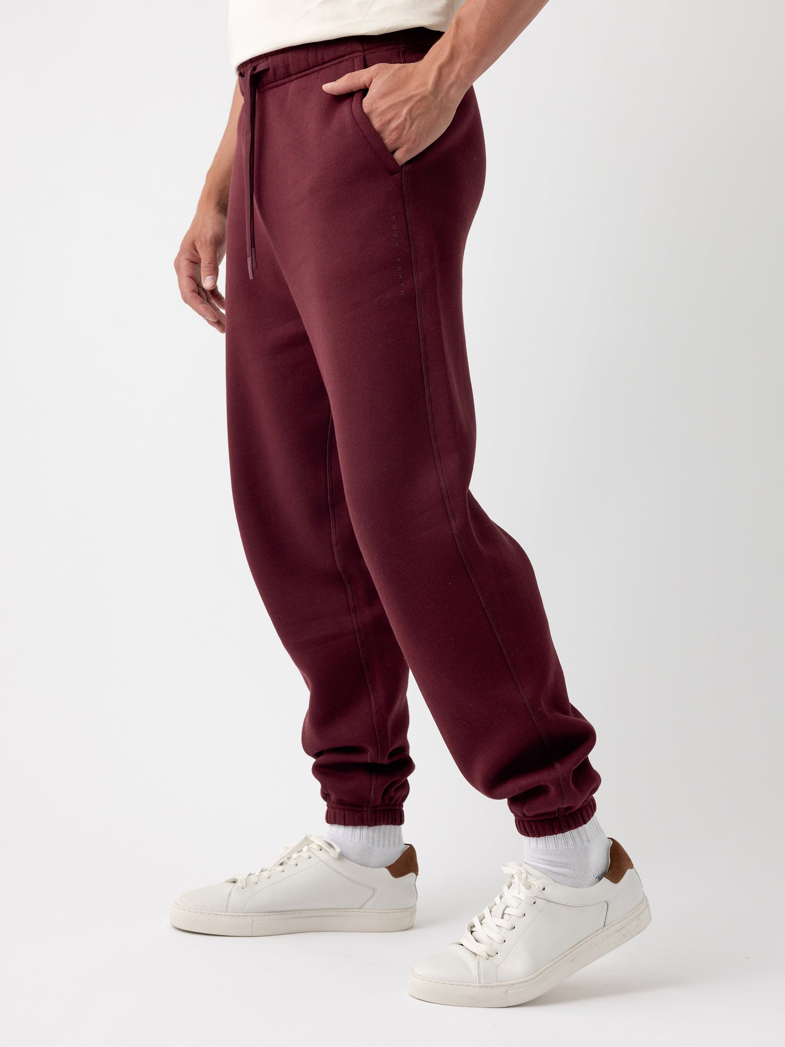 A person is sporting Cozy Earth's Men's CityScape Sweatpant in burgundy, casually resting their hand in the pocket. These sweatpants feature a drawstring at the waist and cuffed ankles. The individual is also wearing white socks and white sneakers with brown accents on the heels. The background is plain white. 