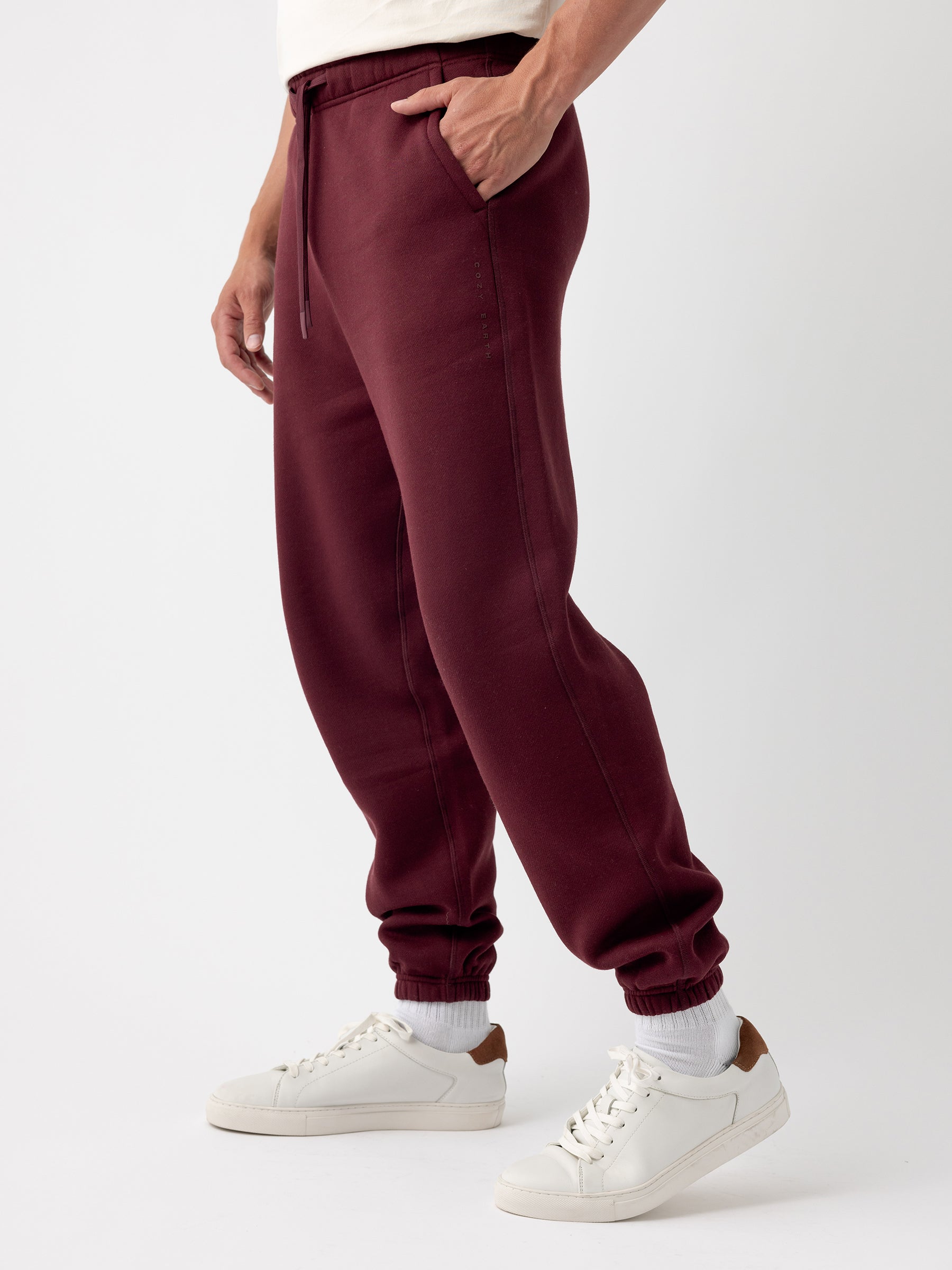 A person is sporting Cozy Earth's Men's CityScape Sweatpant in burgundy, casually resting their hand in the pocket. These sweatpants feature a drawstring at the waist and cuffed ankles. The individual is also wearing white socks and white sneakers with brown accents on the heels. The background is plain white. |Color:Burgundy