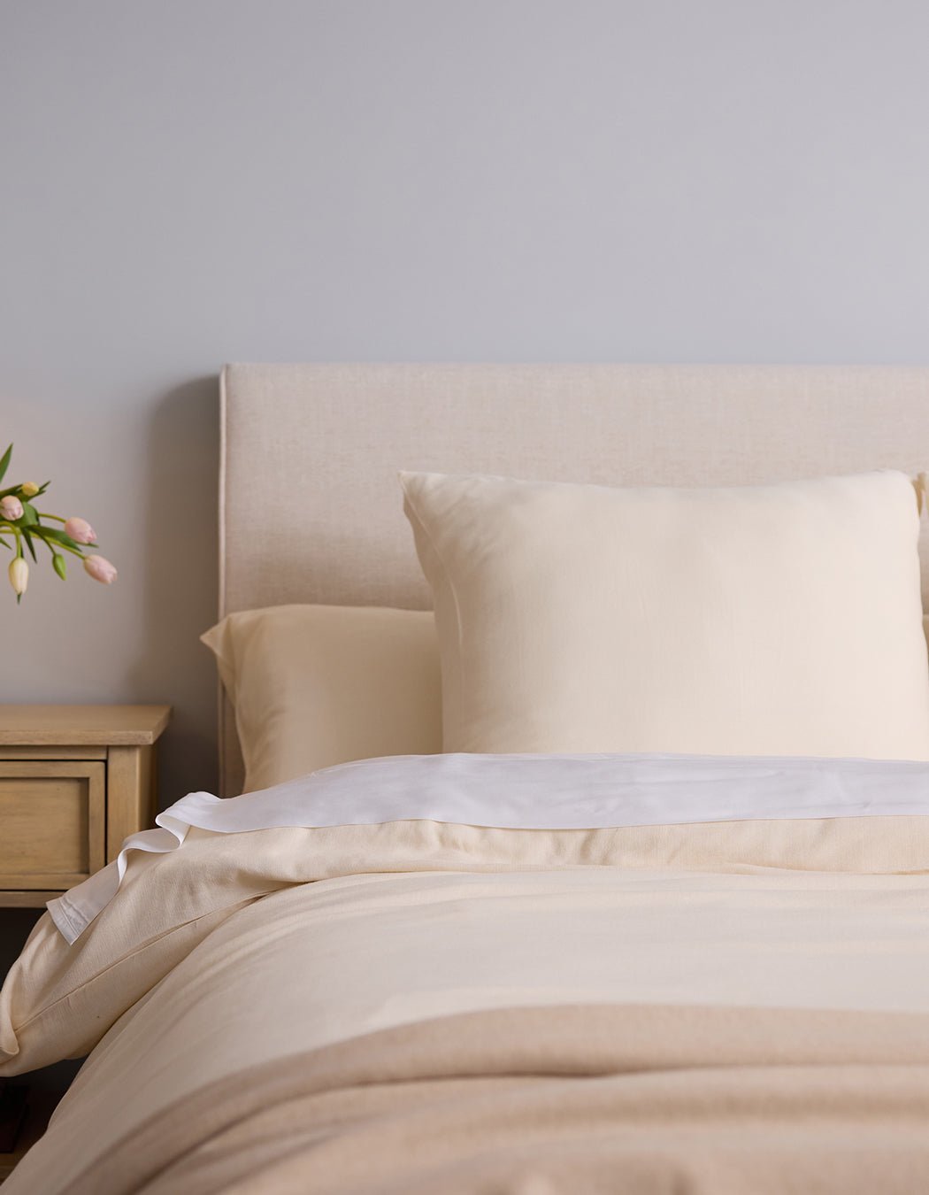 A neatly made bed features Cozy Earth's Aire Bamboo Shams with cream-colored bedding against a light upholstered headboard. On the left, a wooden nightstand holds a vase of pink tulips.