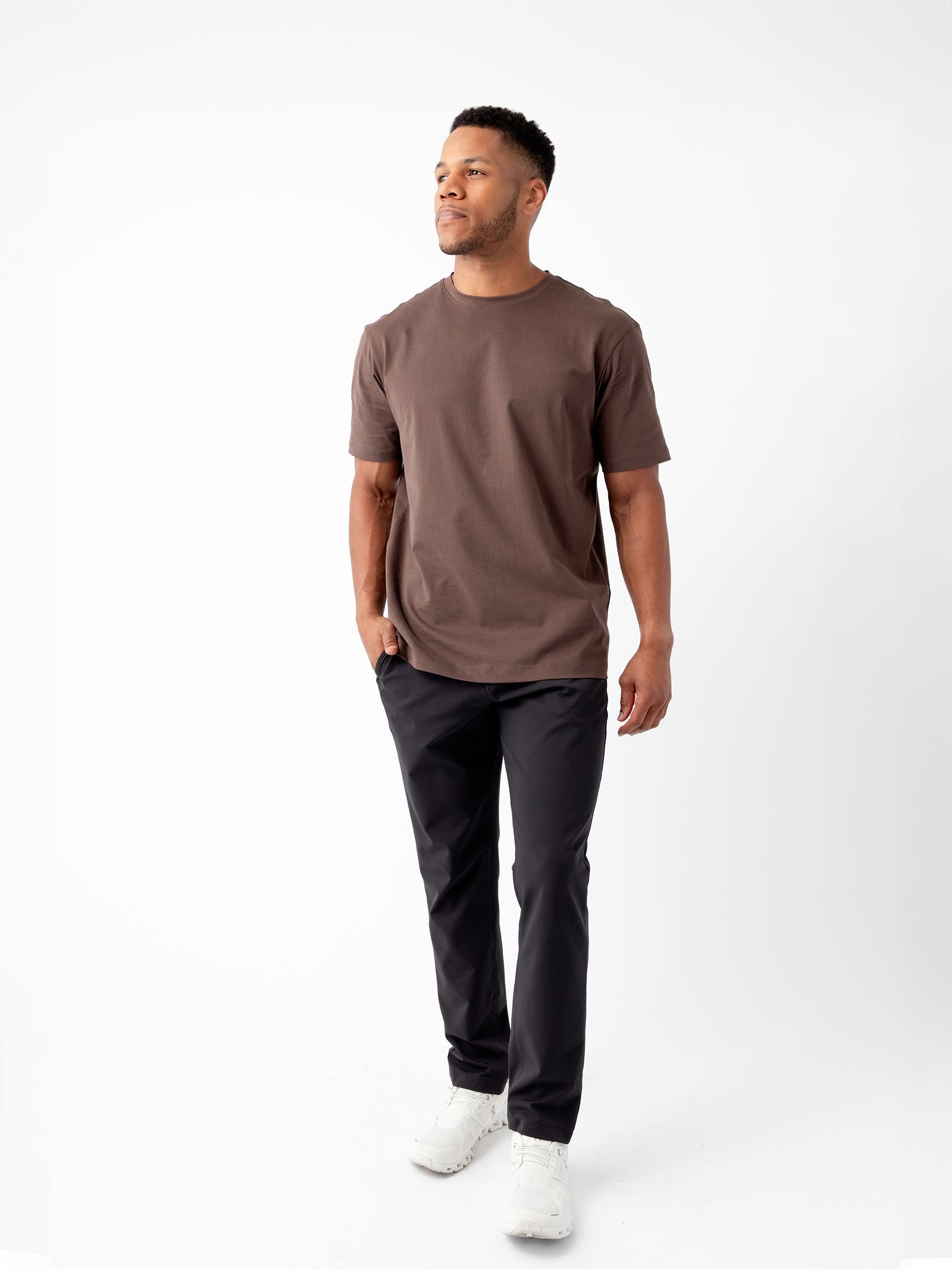 A man stands against a white background, wearing Cozy Earth's brown Men's All Day Tee, black pants, and white sneakers. He gazes slightly to the side with a relaxed posture, one hand in his pocket. 