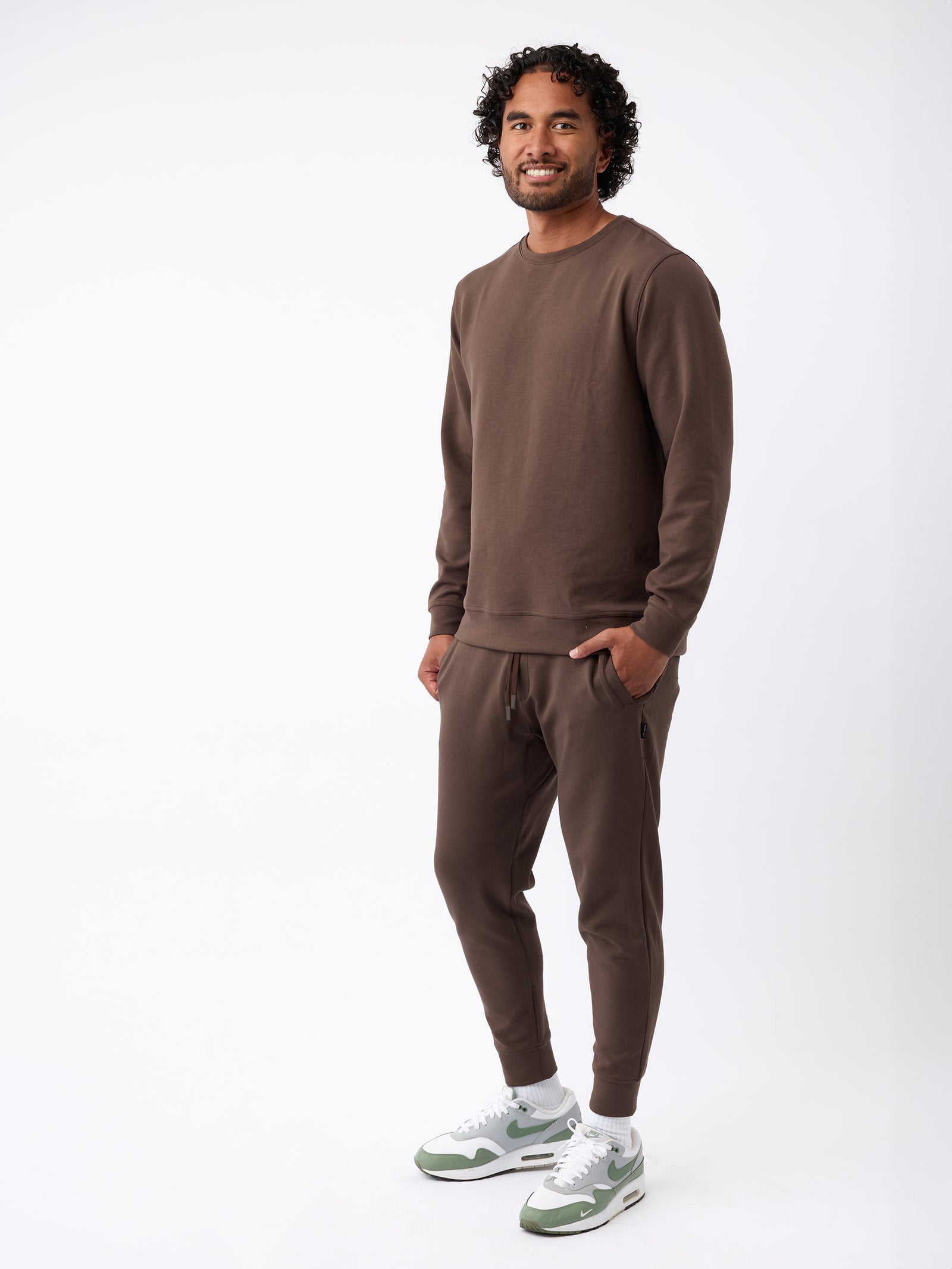 A man with curly hair is smiling and standing against a plain white background. He is wearing Cozy Earth's Men's StretchTech Crewneck and matching joggers in brown, paired with white sneakers featuring green accents. 