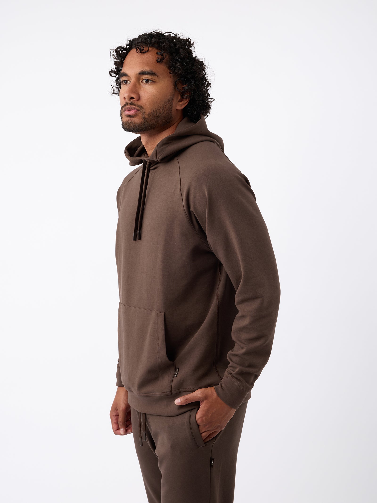 A person with curly hair is standing in profile against a plain white background, wearing the Cozy Earth Men's StretchTech Hoodie and matching brown pants. Their hands are partially in the front pockets of the hoodie. 