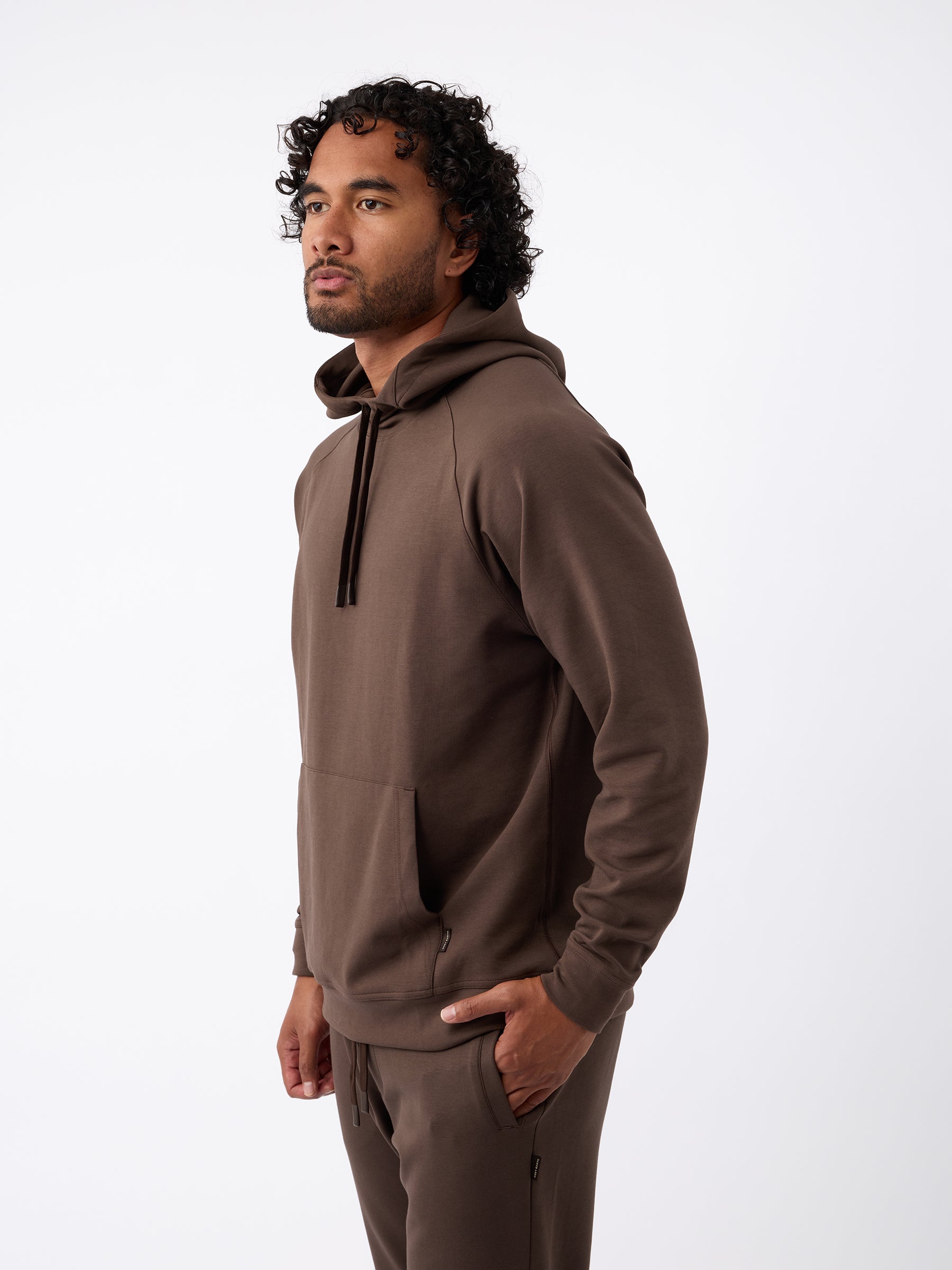 A person with curly hair is standing in profile against a plain white background, wearing the Cozy Earth Men's StretchTech Hoodie and matching brown pants. Their hands are partially in the front pockets of the hoodie. |Color:Cacao