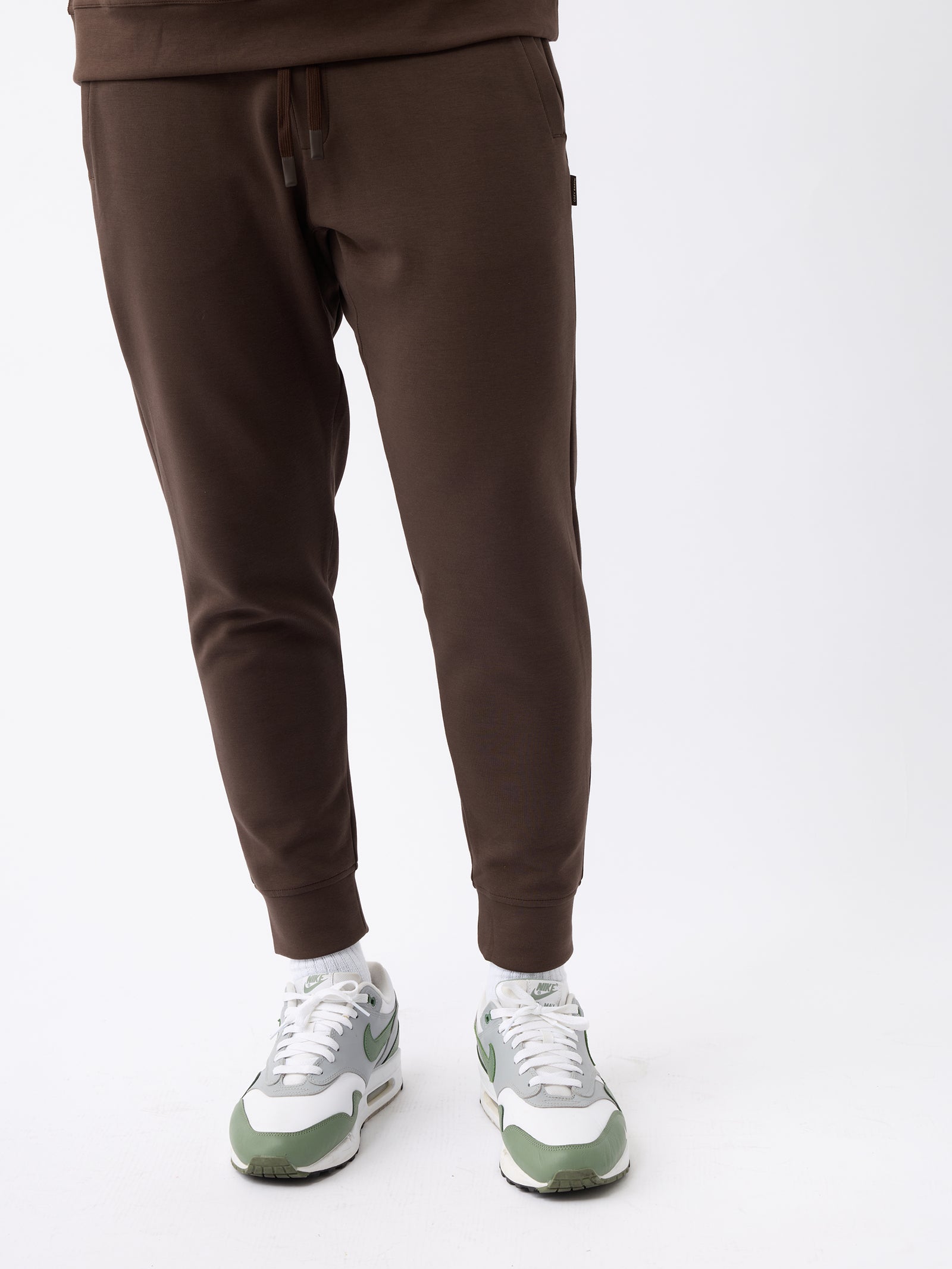 A person wearing Cozy Earth's Men's StretchTech Jogger in chocolate brown and white and green sneakers stands against a plain white background. The image focuses on the lower half of the body, highlighting the casual attire. 