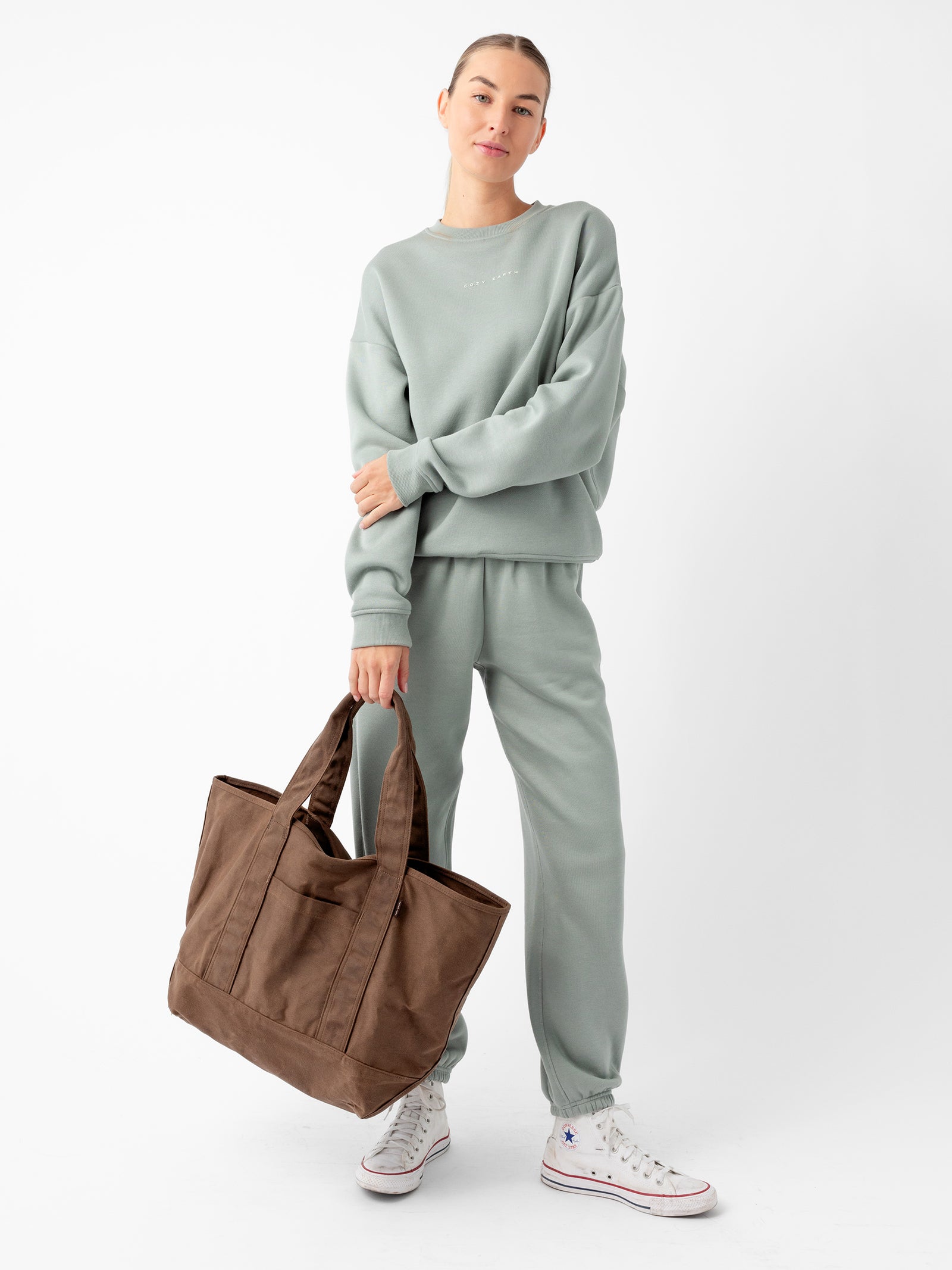 A person wearing a pastel green sweatshirt and matching sweatpants stands against a plain white background. They are carrying a large Waxed Canvas Tote from Cozy Earth and wearing white sneakers. They have light-colored hair pulled back in a neat style and are smiling slightly. 