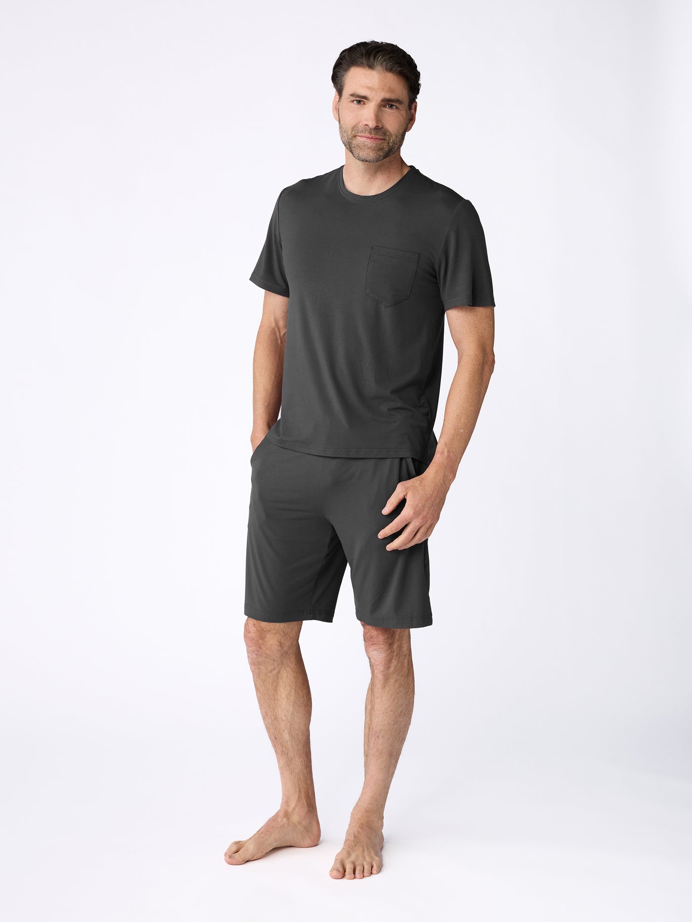 A man stands barefoot against a plain white background, wearing the Cozy Earth Men's Bamboo Stretch-Knit Short Sleeve Pajama Set in dark gray, featuring a chest pocket t-shirt and knee-length shorts. |Color:Charcoal