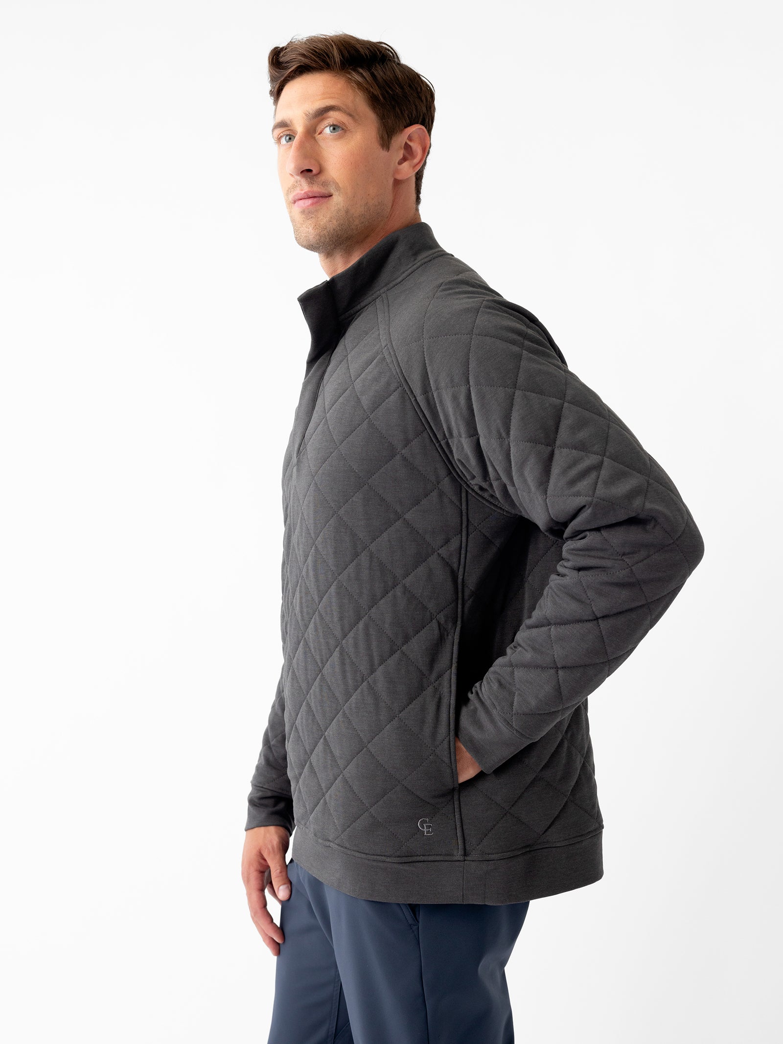 A man wearing the Men's Ultra-Soft Bamboo Quilted Snap Pullover in dark gray by Cozy Earth and dark blue pants stands against a plain white background. He is posing with both hands in his pockets and looking slightly to his left. The pullover features a high collar and a quilted pattern. 