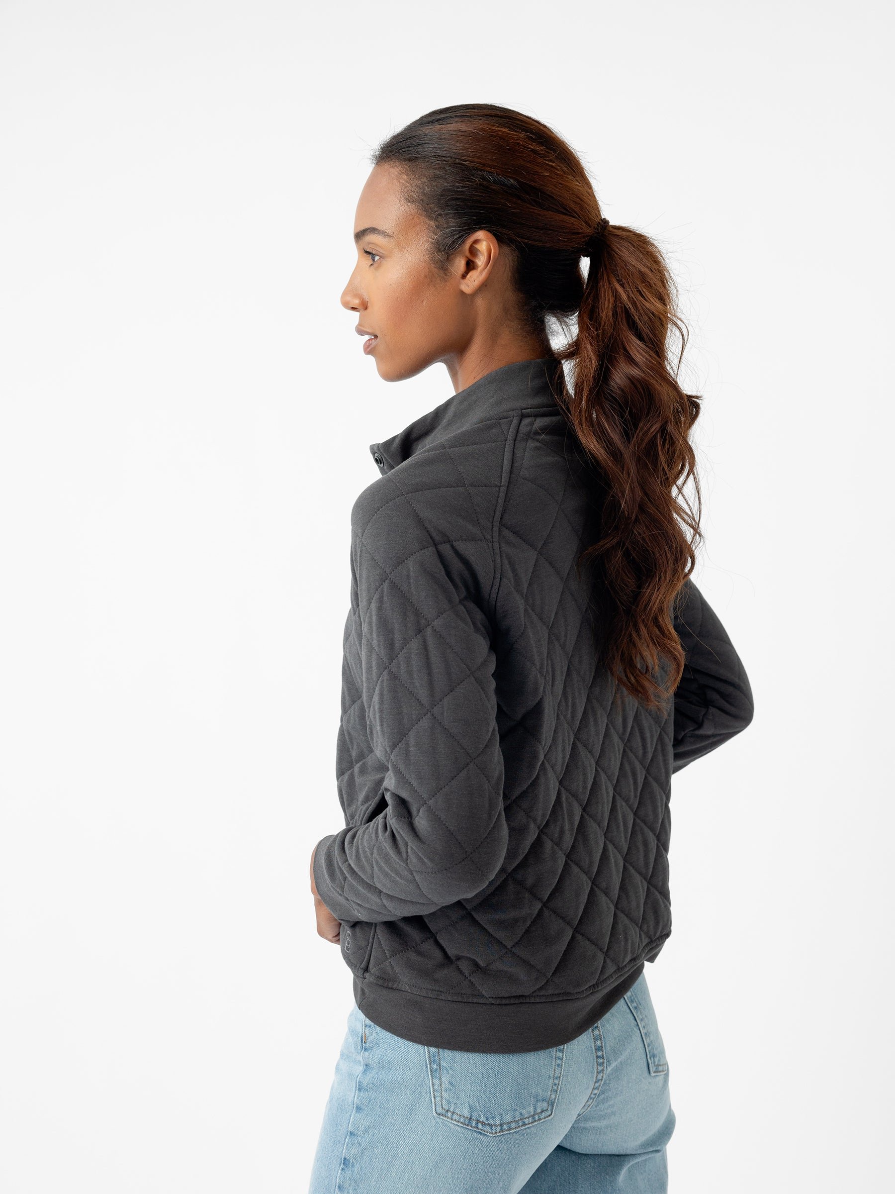 A person with long hair styled in a ponytail is turned to the side, dressed in Cozy Earth's Women's Ultra-Soft Bamboo Quilted Snap Pullover and light blue jeans. The background is white and minimalistic. |Color:Charcoal