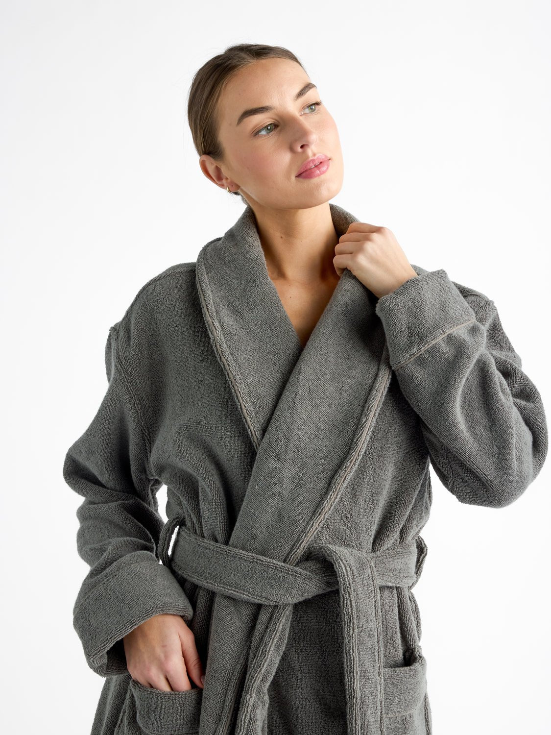 A woman models the Luxe Bath Robe by Cozy Earth, featuring charcoal fabric. They are pictured from the waist up and gazing to the side while gently holding the robe's collar. 