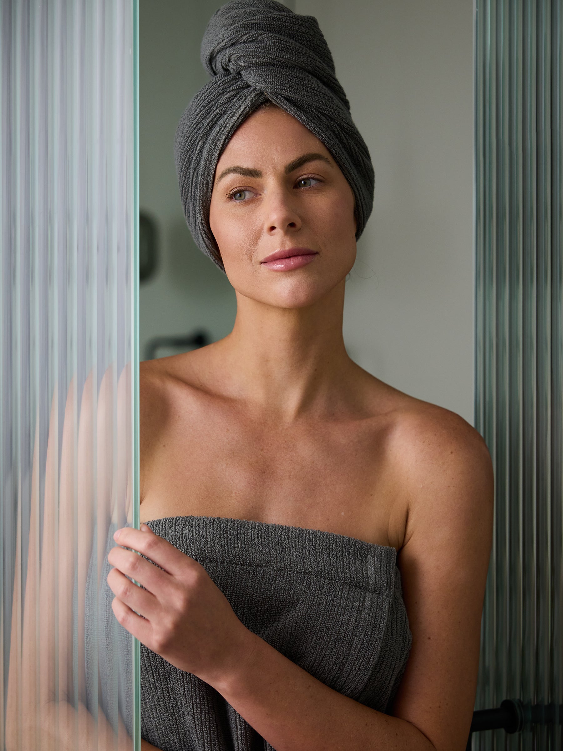 A woman with a Cozy Earth Ribbed Terry Hair Towel wrapped around her head and another grey towel around her body stands next to a frosted glass shower door, looking off into the distance. She has a calm and relaxed expression on her face. |Color:Charcoal