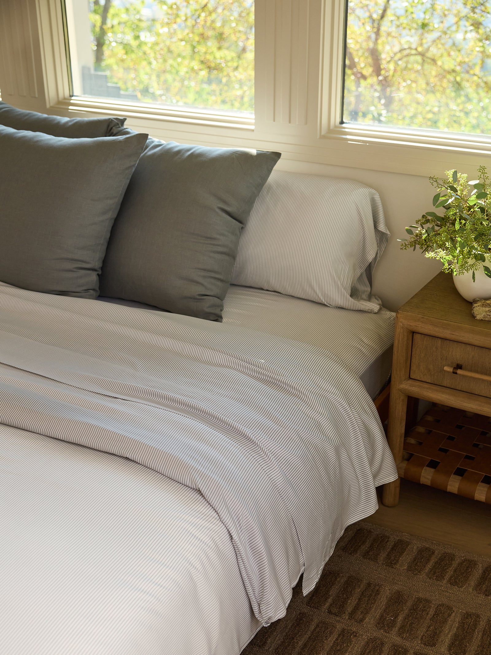 A neatly made bed showcases the Cozy Earth Bamboo Duvet Cover, designed with white and gray stripes, complemented by three gray pillows. A wooden nightstand holds a small plant, while sunlight streams through the windows, casting a warm glow on the bed. A brown woven rug is partially visible on the floor. 