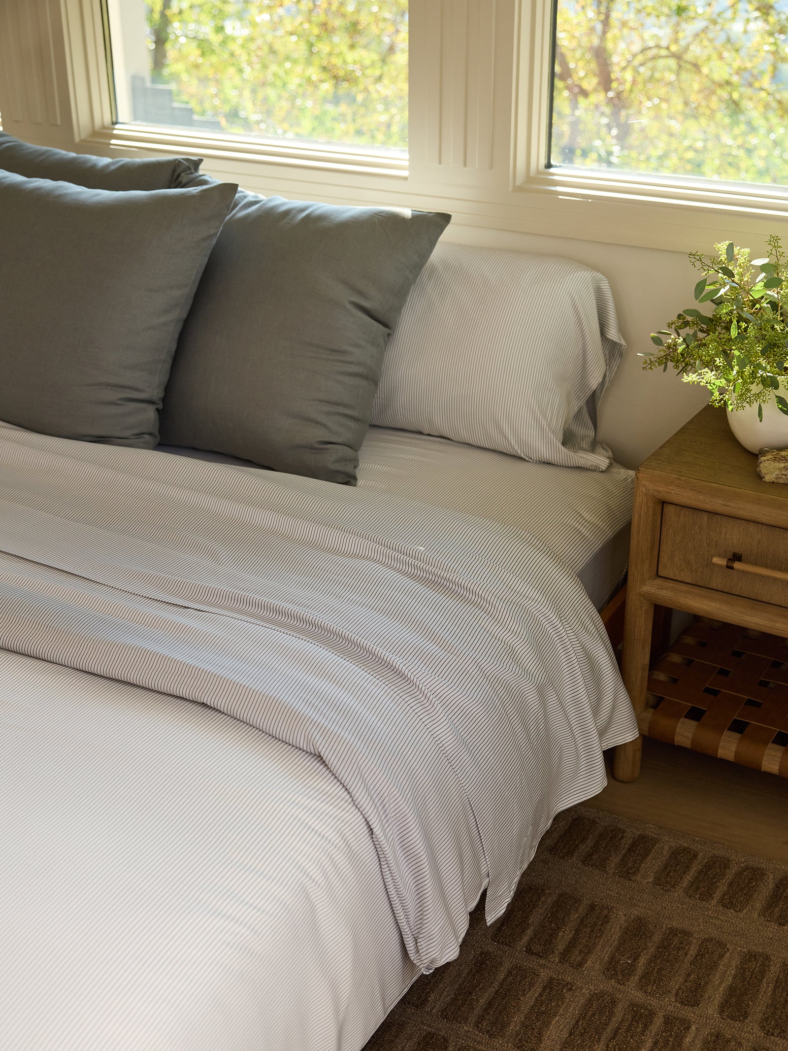 A cozy bedroom scene showcases a neatly made bed adorned with the Bamboo Flat Sheet from Cozy Earth, featuring white and gray stripes and complemented by three gray pillows. A wooden nightstand beside the bed holds a small plant, while sunlight streams through the windows, casting a warm glow. 