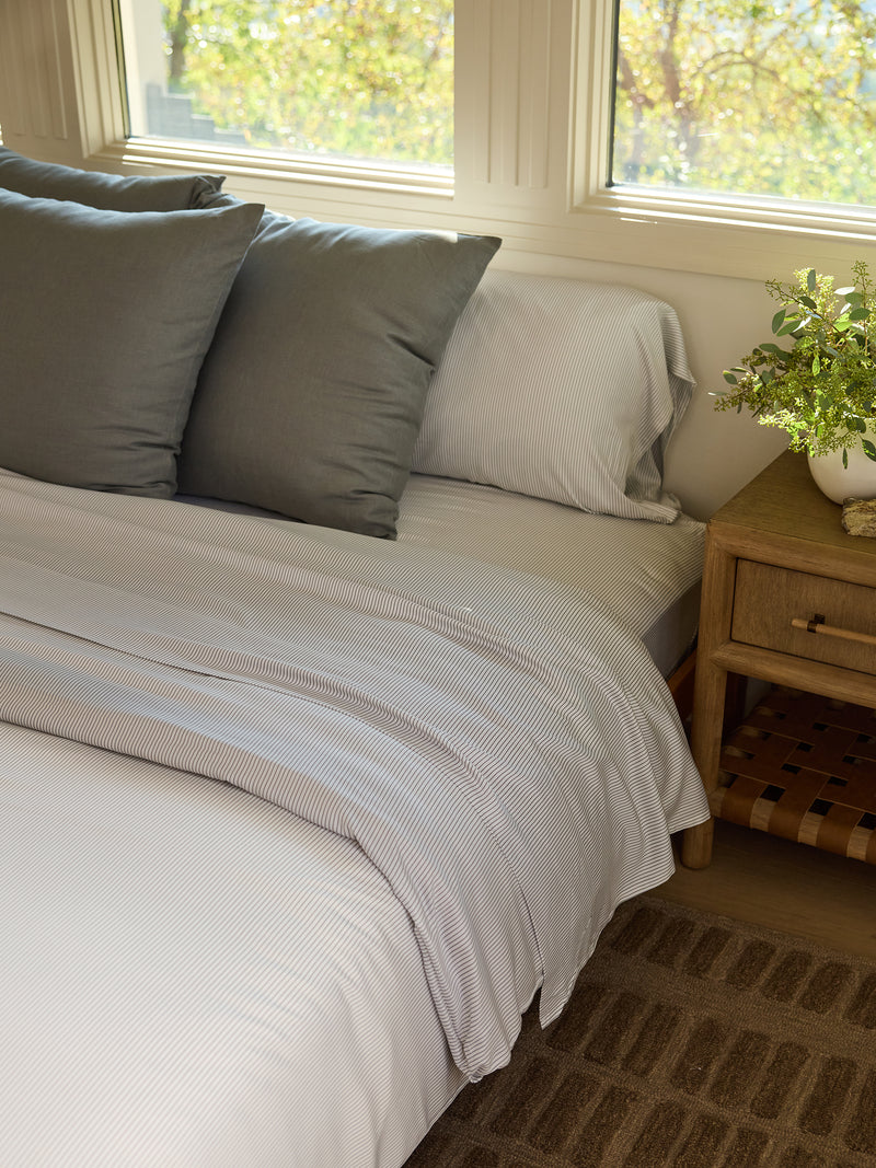 A cozy bedroom scene showcases a neatly made bed adorned with the Bamboo Flat Sheet from Cozy Earth, featuring white and gray stripes and complemented by three gray pillows. A wooden nightstand beside the bed holds a small plant, while sunlight streams through the windows, casting a warm glow.
