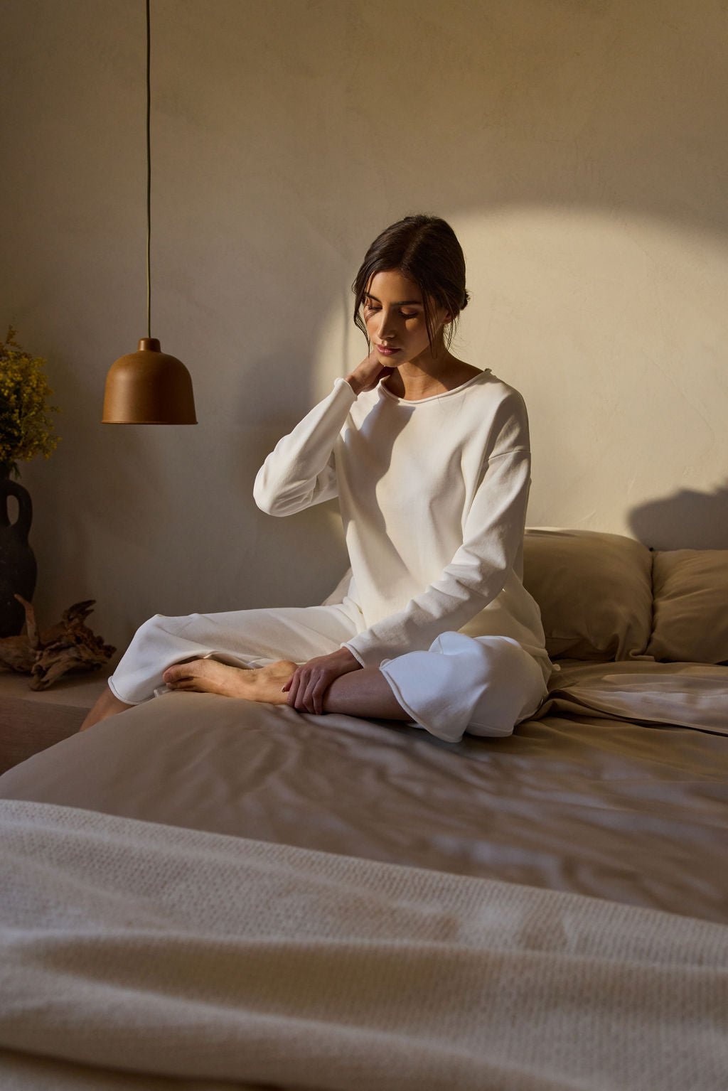 In a softly lit room, a person wearing Cozy Earth's Women's Chelsea Cropped Pant sits on a bed. A brown pendant light hangs above, with a vase of flowers in the background, creating a serene ambiance. 