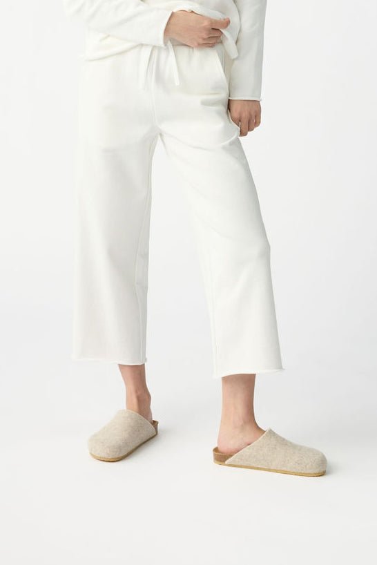 A person in Cozy Earth's Women's Chelsea Set, featuring loose-fitting white pants and a long-sleeve top, stands against a plain white background, paired with minimalist off-white slip-on shoes. 