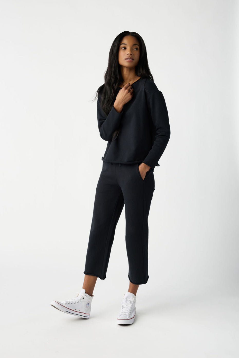 A person with long hair stands against a plain backdrop wearing Cozy Earth's Women's Chelsea Set, paired with white high-top sneakers. One hand is in their pocket while the other is held near their chest. |Color:Jet Black