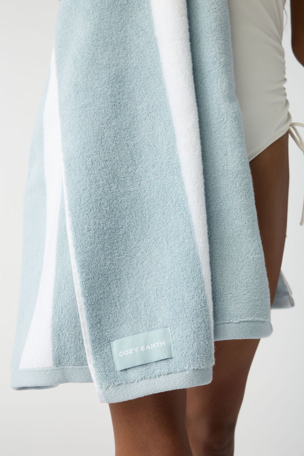 A person in a white swimsuit is draped with a Classic Stripe Resort Towel, displaying the "Cozy Earth" label, in light blue and white stripes. 