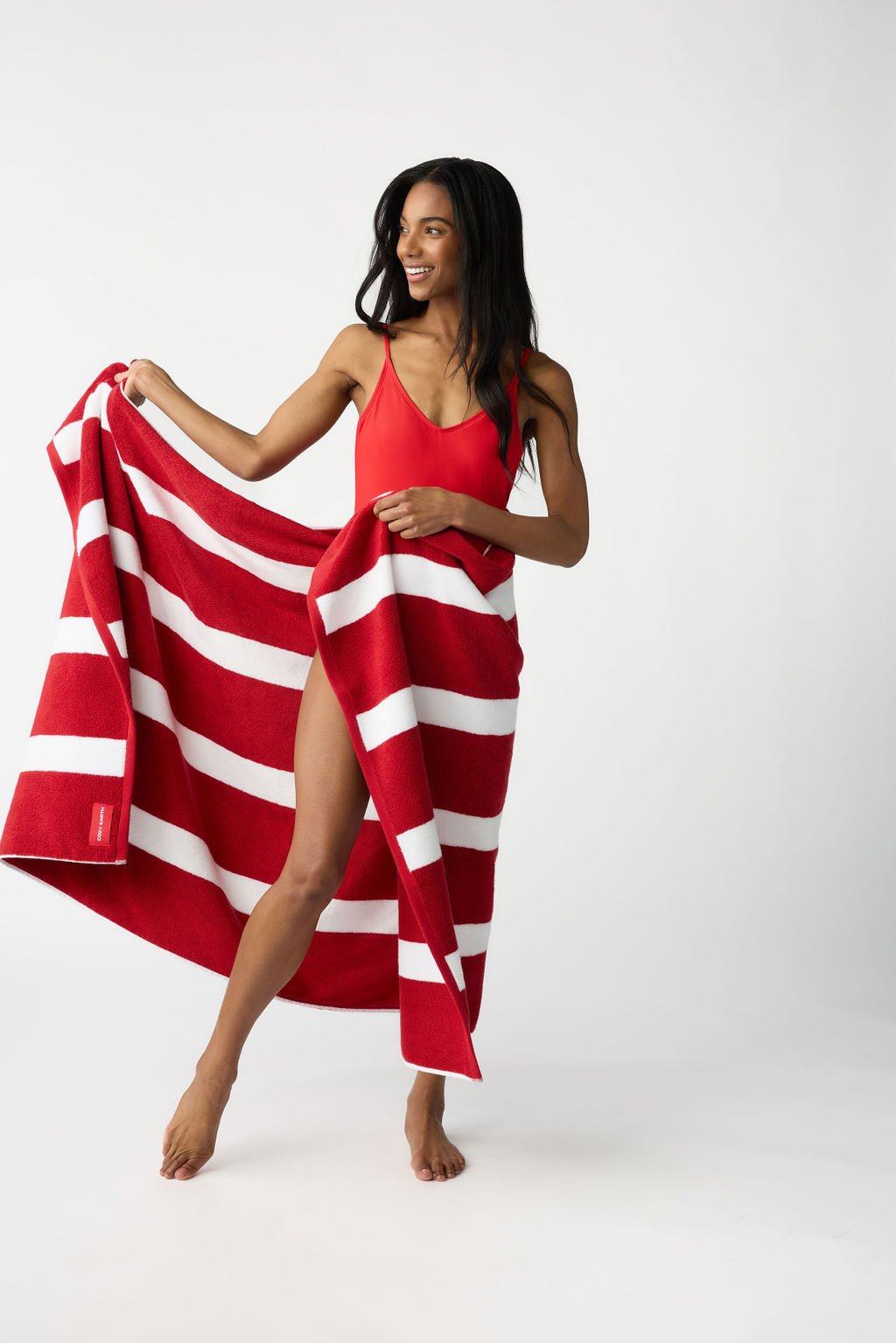 A woman in a red swimsuit holds the Cozy Earth Classic Stripe Resort Towel, dressed in striking red and white stripes, partially wrapped around her. She stands barefoot against a plain white backdrop, smiling and looking to her right. 