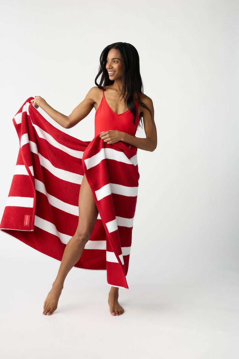A woman in a red swimsuit holds the Cozy Earth Classic Stripe Resort Towel, dressed in striking red and white stripes, partially wrapped around her. She stands barefoot against a plain white backdrop, smiling and looking to her right.