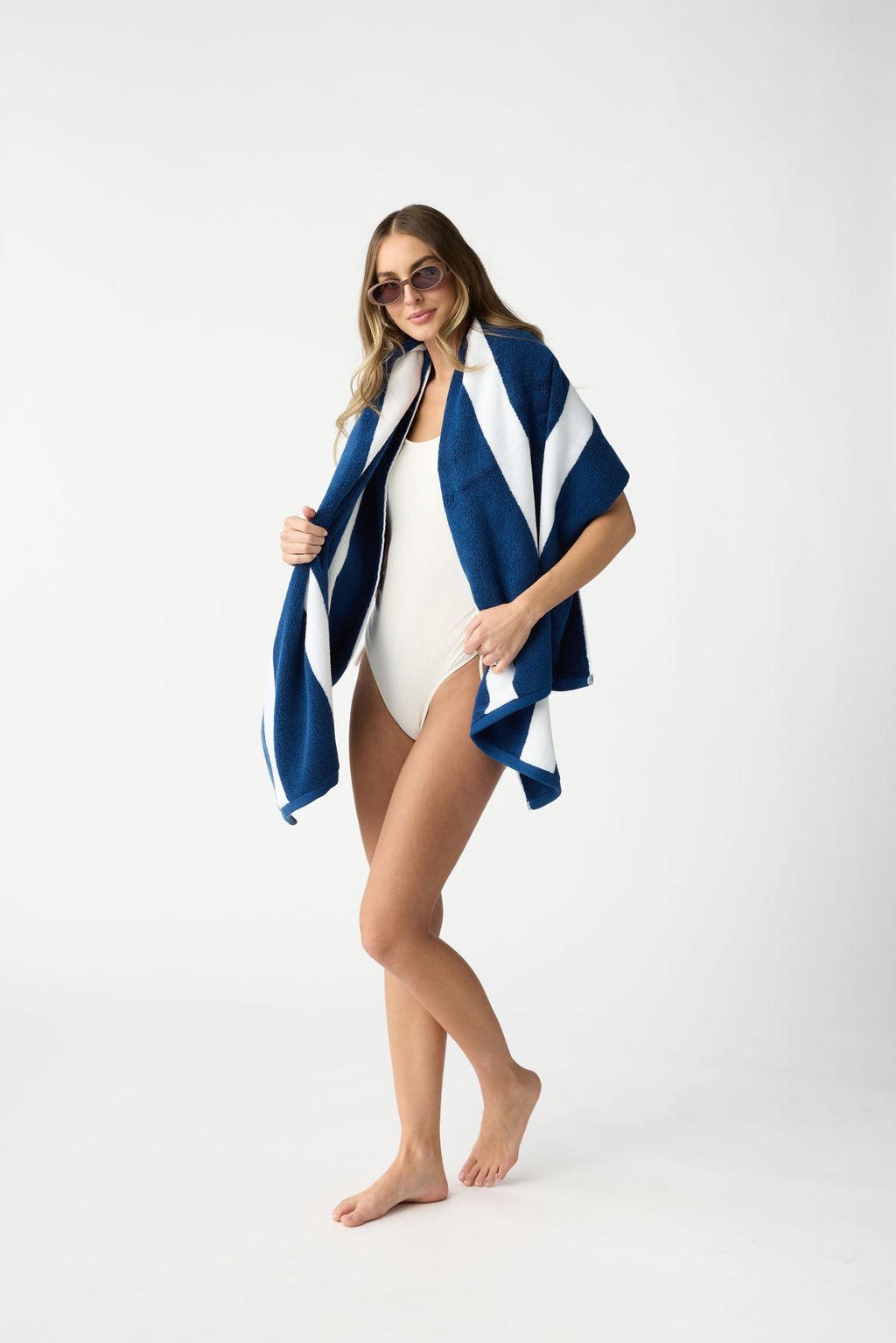 A woman in sunglasses and a white swimsuit stands barefoot, smiling while draped with a Cozy Earth Classic Stripe Resort Towel against a plain white background. |Color:Pacific