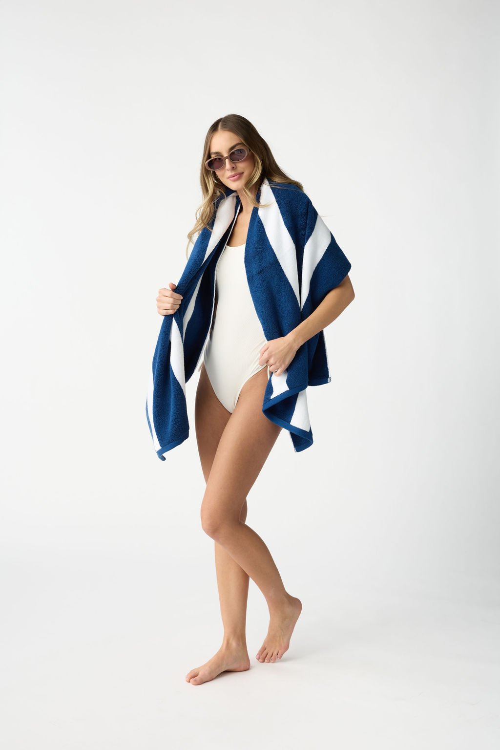 A woman in sunglasses and a white swimsuit stands barefoot, smiling while draped with a Cozy Earth Classic Stripe Resort Towel against a plain white background. |Color:Pacific