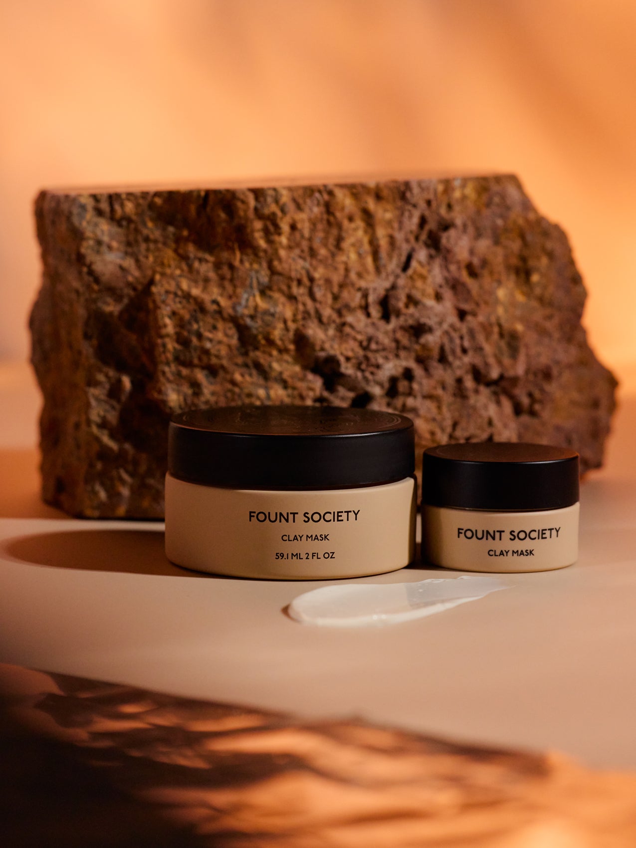 Two jars of Cozy Earth's Clay Mask sit on a beige surface with a textural brown rock backdrop. A clay mask smear is visible in front, highlighting a natural, earthy theme. 