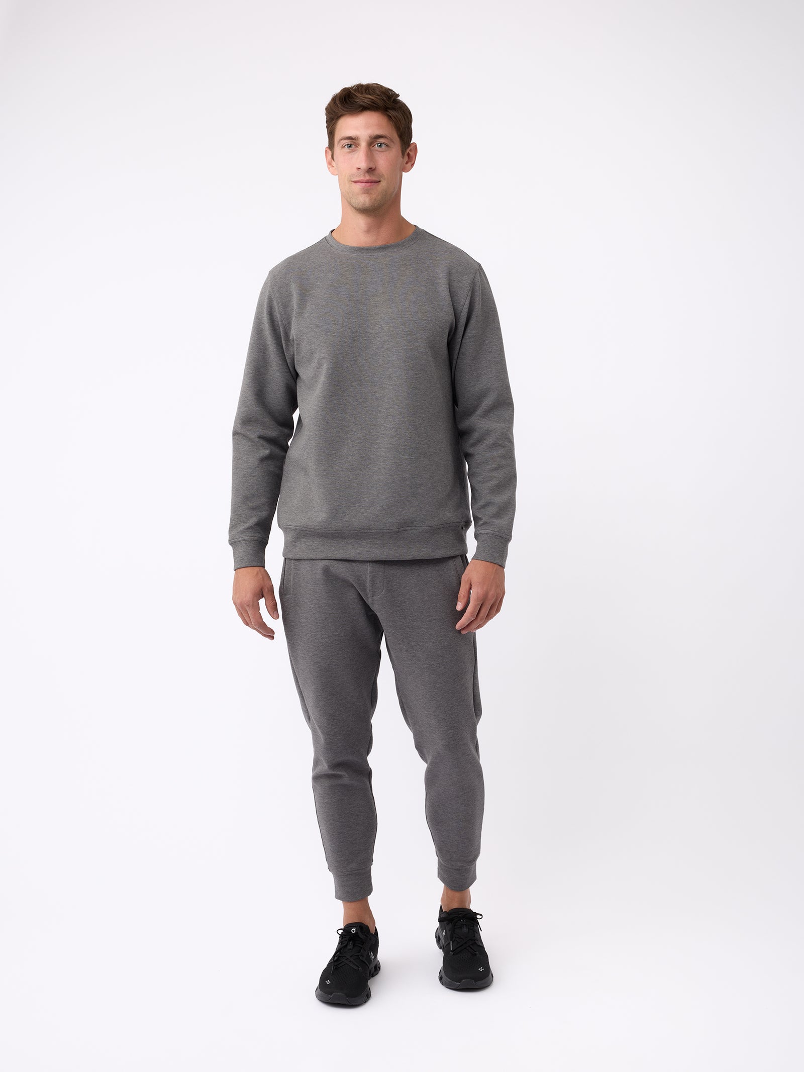 A person dressed in Cozy Earth's Men's StretchTech Jogger, featuring a gray sweatshirt and matching sweatpants, stands against a plain white background. They have short brown hair and are sporting black sneakers. The outfit exudes a casual and comfortable vibe. 