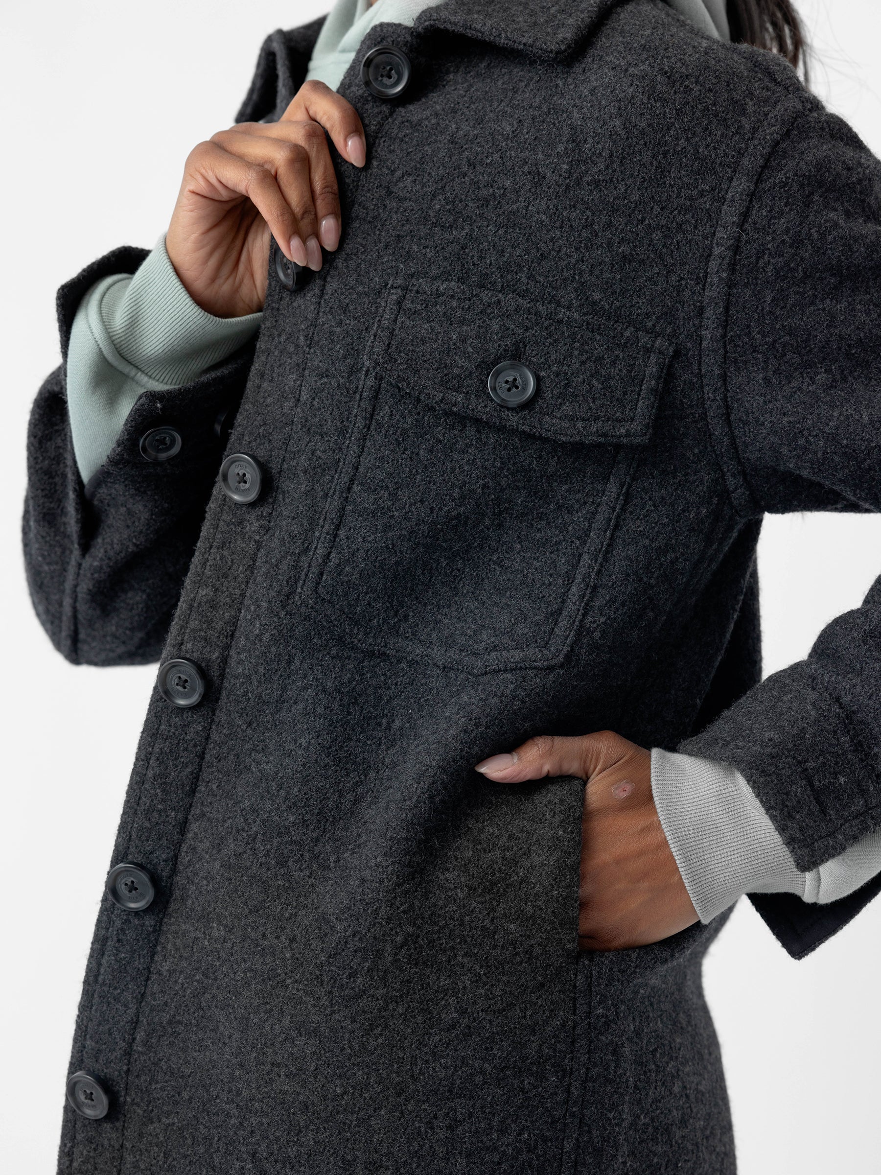 A person is shown from the neck down, wearing the Women's Boucle Shacket by Cozy Earth in dark gray, featuring a breast pocket and a glimpse of a light gray sleeve underneath. One hand grasps the shacket's collar while the other is tucked into a side pocket. The background is plain and white. |Color:Coal