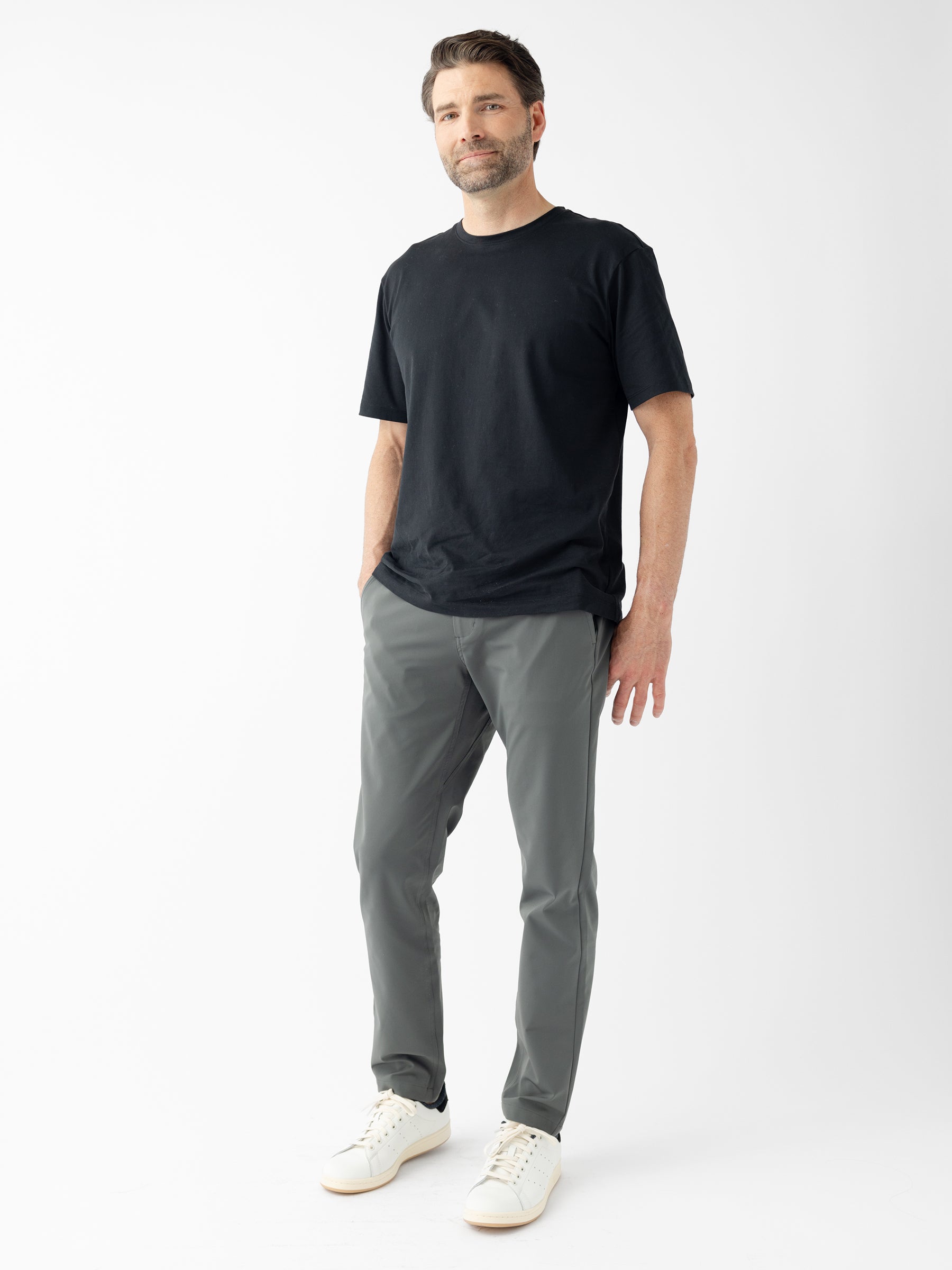 A man stands against a plain white background, casually dressed in a black t-shirt, Cozy Earth's Men's Everywhere Pant 30L in gray, and white sneakers. With his hands in his pockets, short dark hair, and a beard, he looks slightly off to the side with a neutral expression. |Color:Coal
